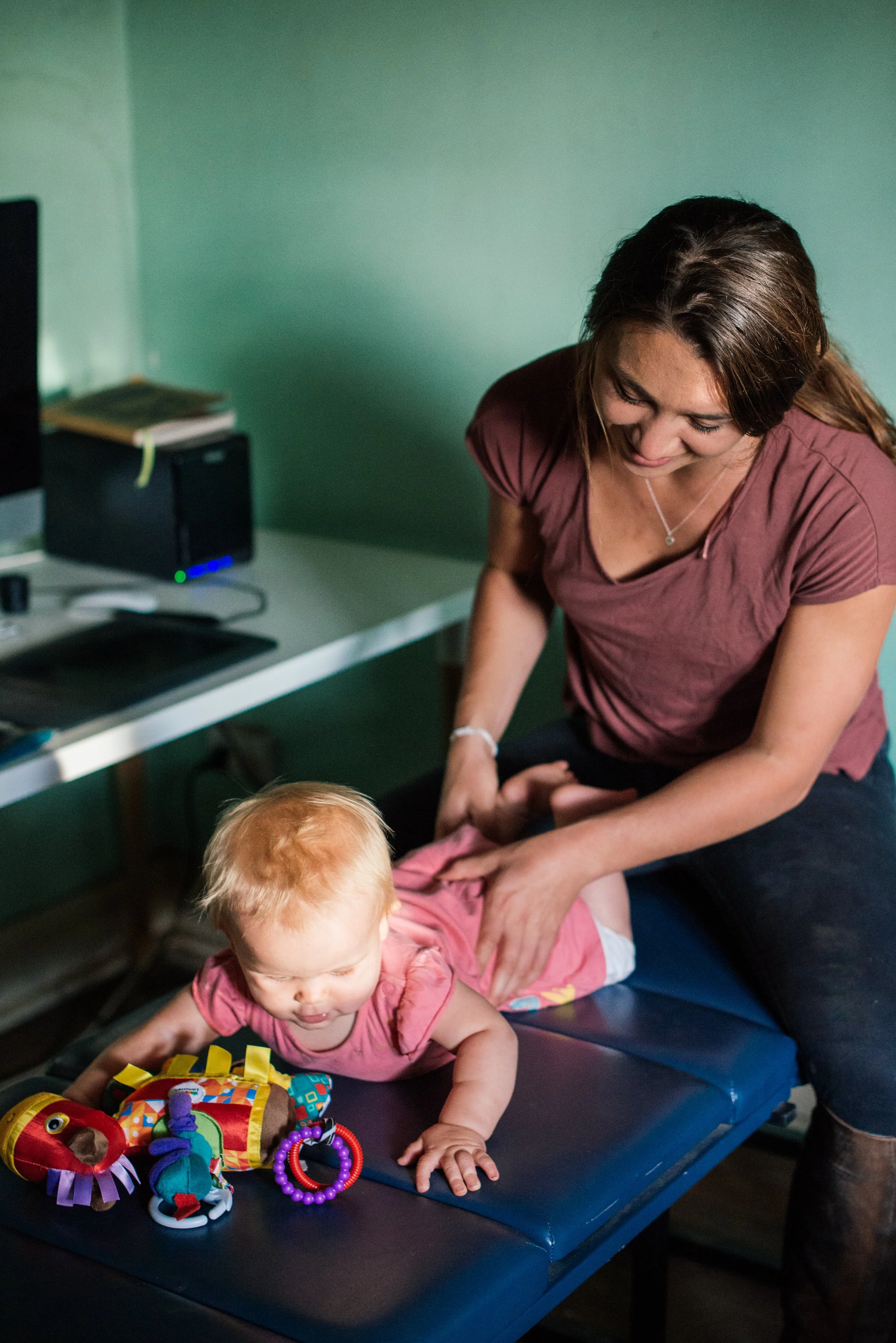 A woman leaning over a young child lying on a padded surface, with a colorful toy in front of the child. The woman is smiling and appears to be interacting with the child. The scene is set in a room with a computer and books on a desk in the background.