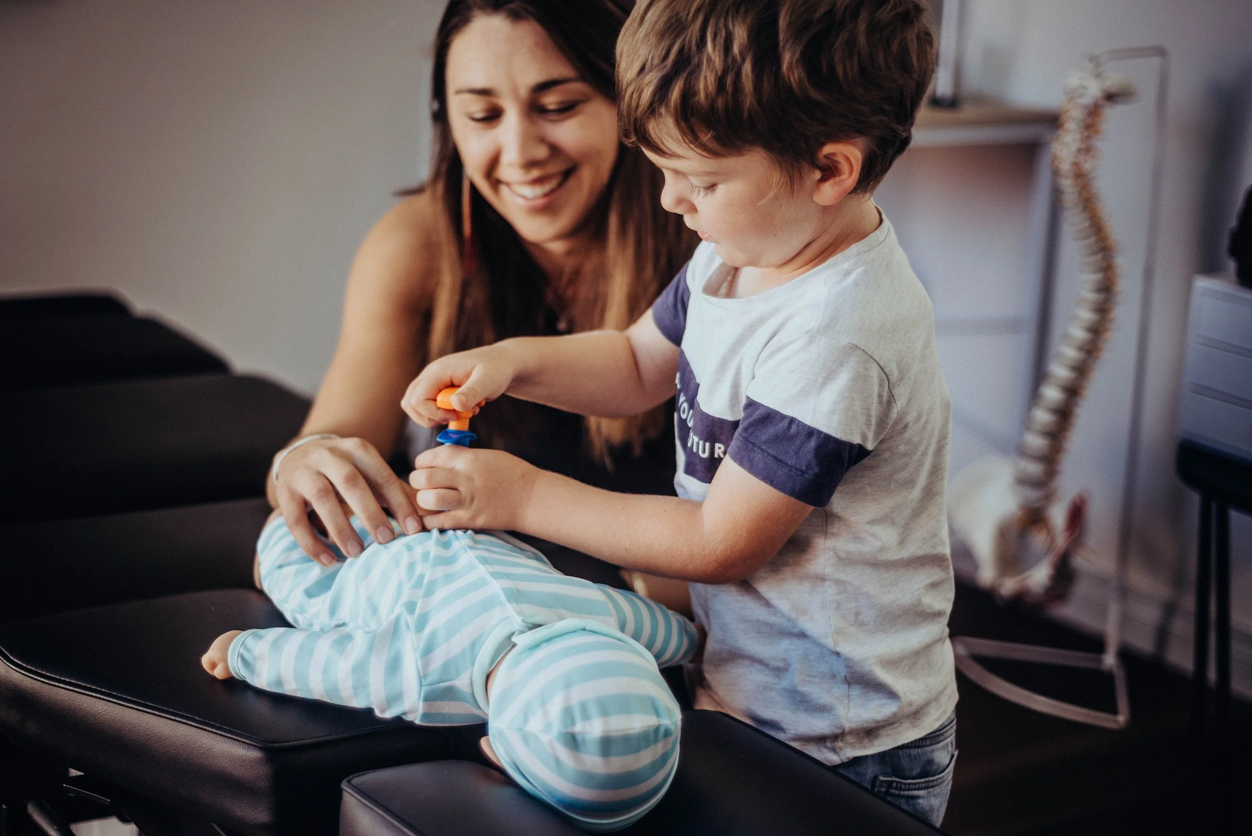 A female medical professional with long dark hair smiling as she assists a young boy with short brown hair during a medical examination or procedure. The boy is lying on an examination table wearing striped pajamas, and he appears focused on the task. The setting is a health clinic or doctor's office.