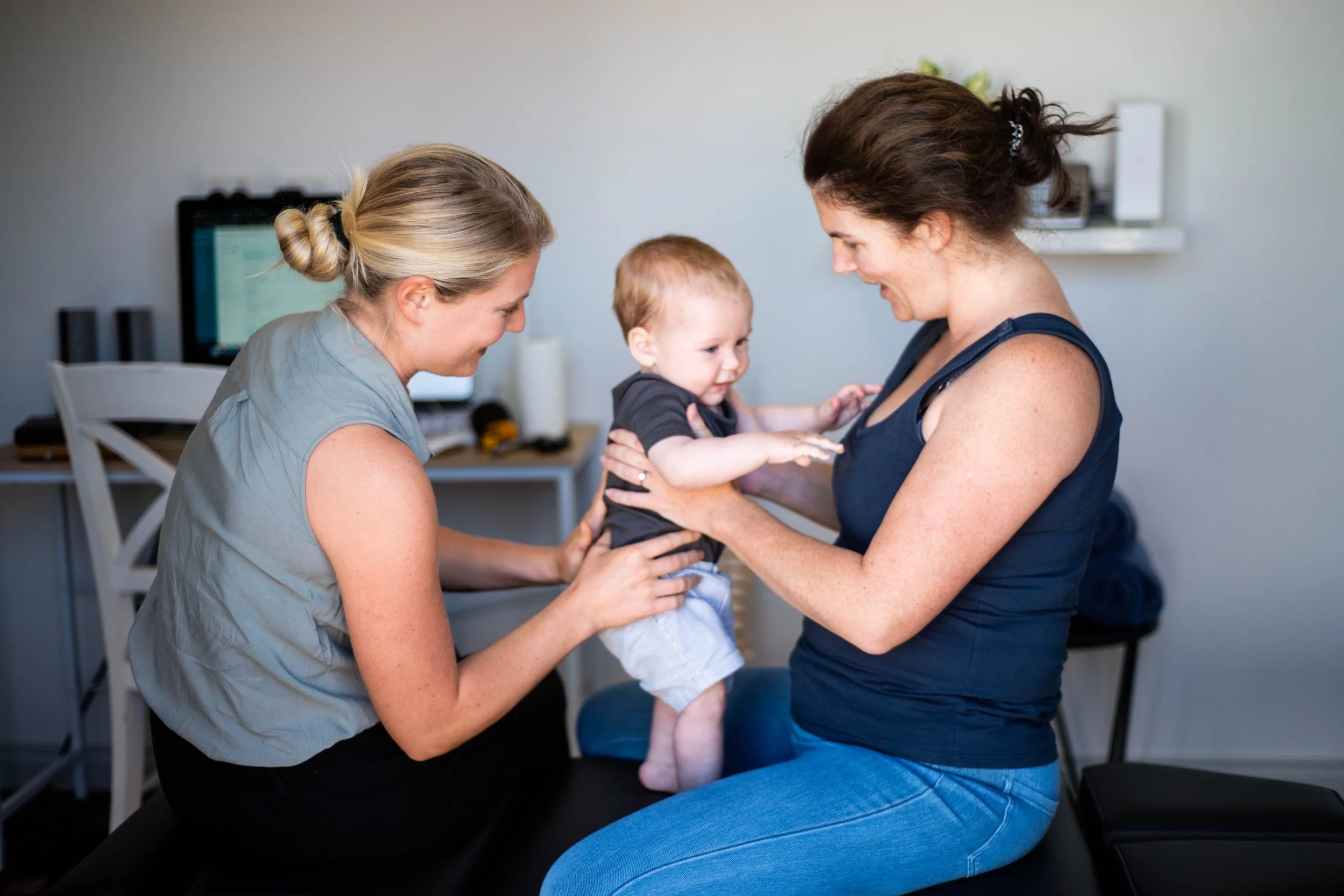 Two women smiling and holding a young child in an indoor setting, with a computer monitor and desk in the background.