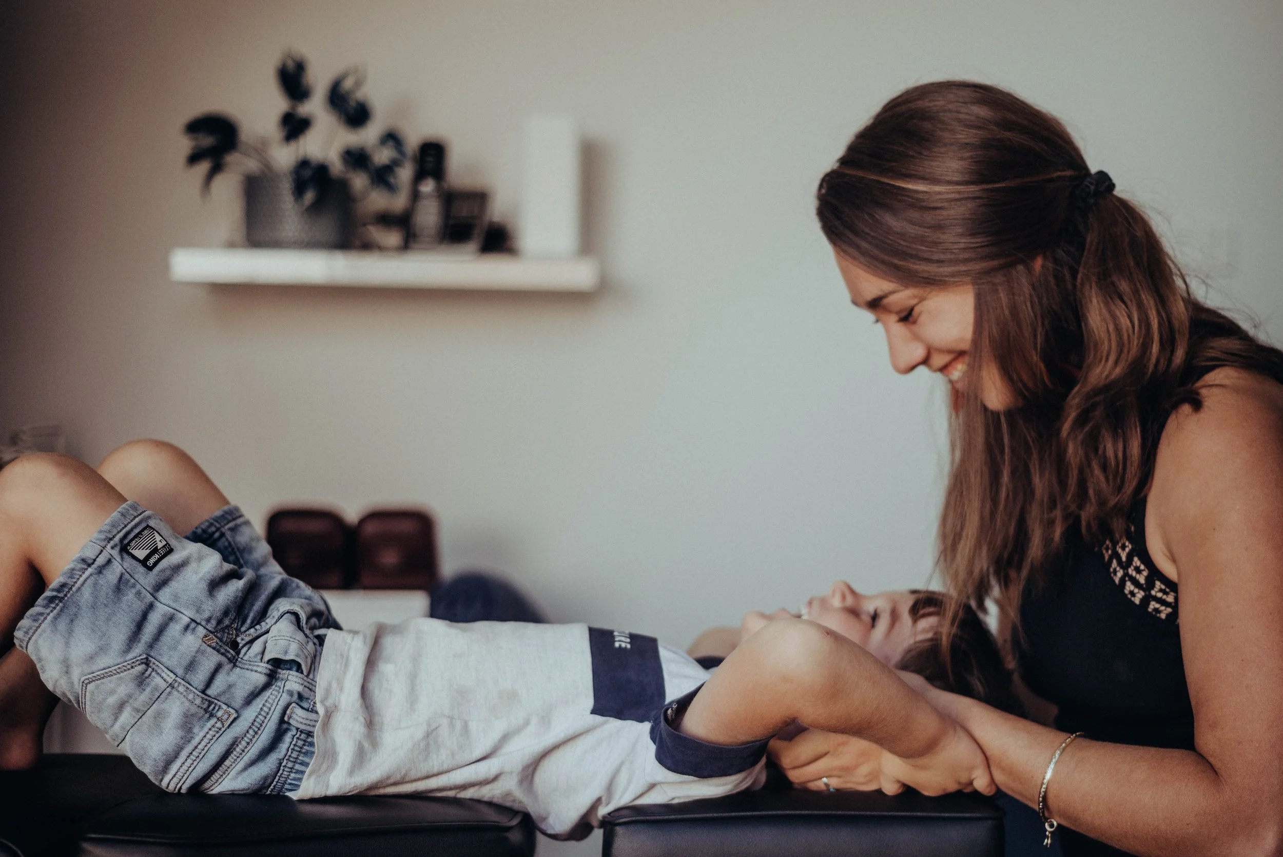 A woman with long brown hair smiling and leaning over a young boy who is lying on a medical examination table with his eyes closed.