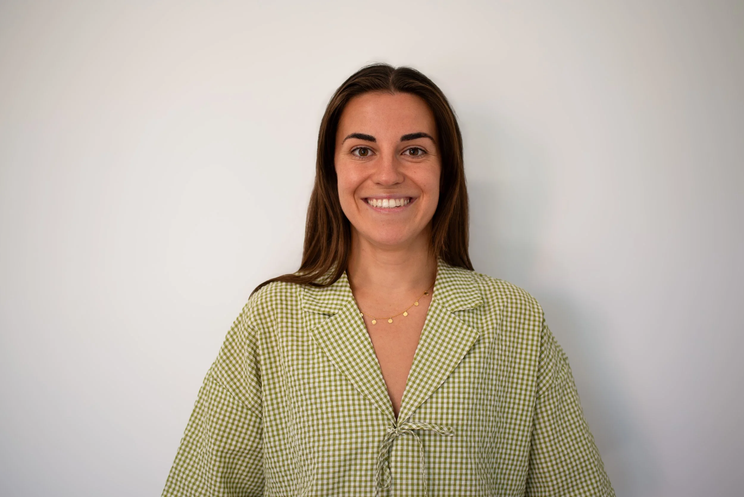 A woman with long brown hair, wearing a green checked shirt with a tied knot, smiling and standing against a plain white wall.