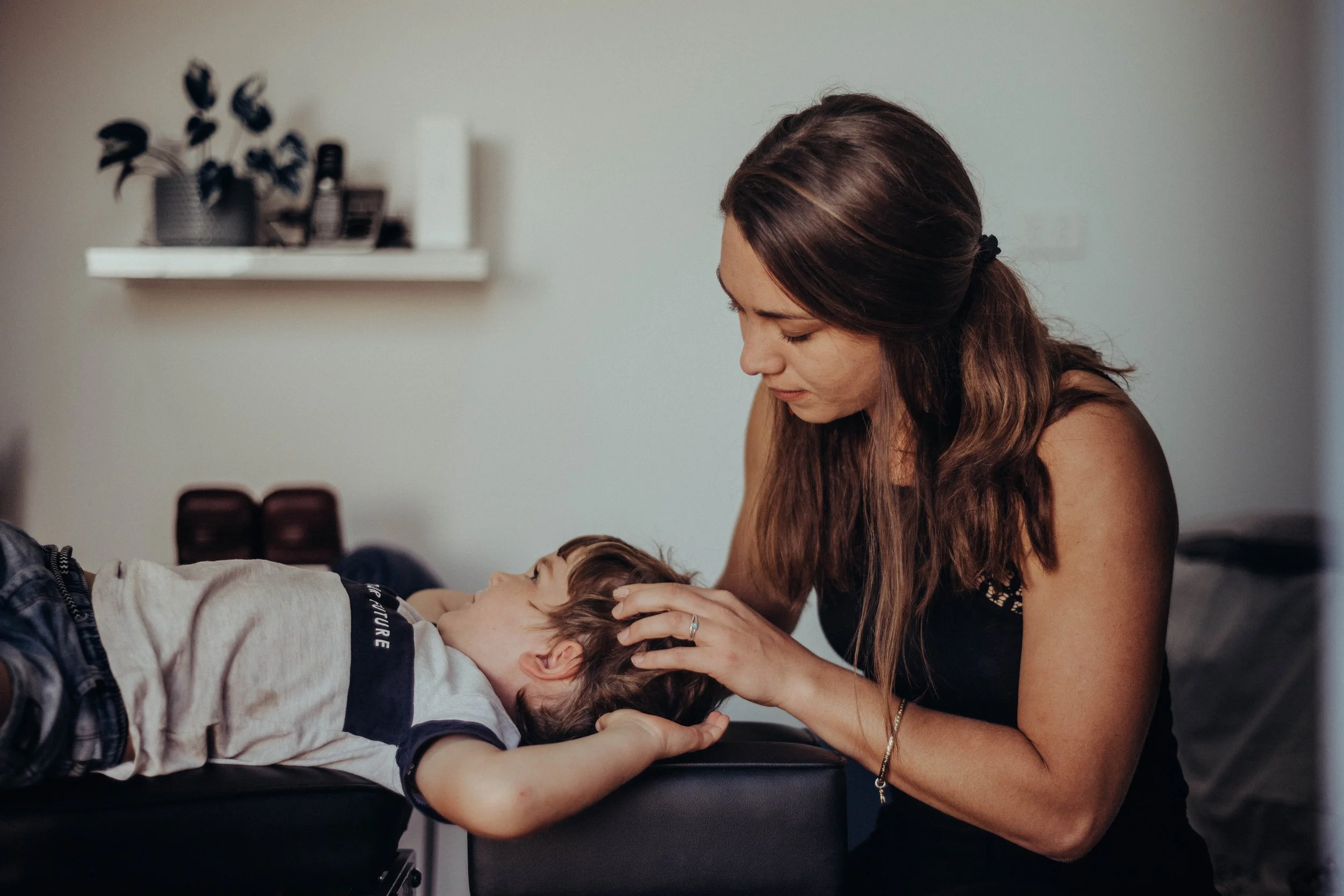 A woman with brown hair pulls back a young boy's hair as he lies down on a black table, with his eyes closed, in a home setting. The woman looks down at him tenderly. In the background, there is a white wall with a floating shelf holding a potted plant and some books.