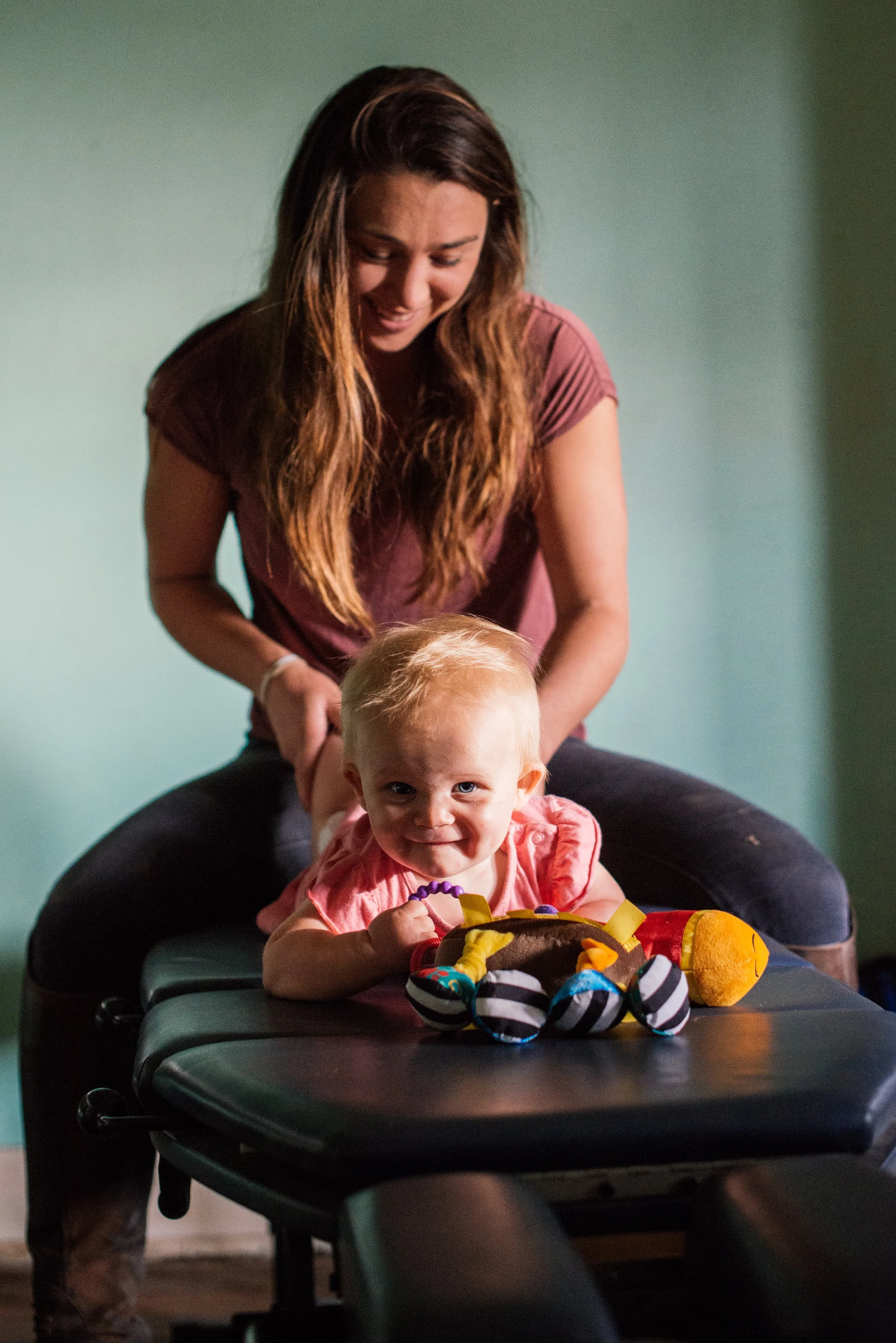 A woman with long brown hair smiling and looking down at a young girl with blond hair sitting on a massage table. The girl is smiling at the camera, holding toys and surrounded by stuffed animals, with a dark green walls in the background.