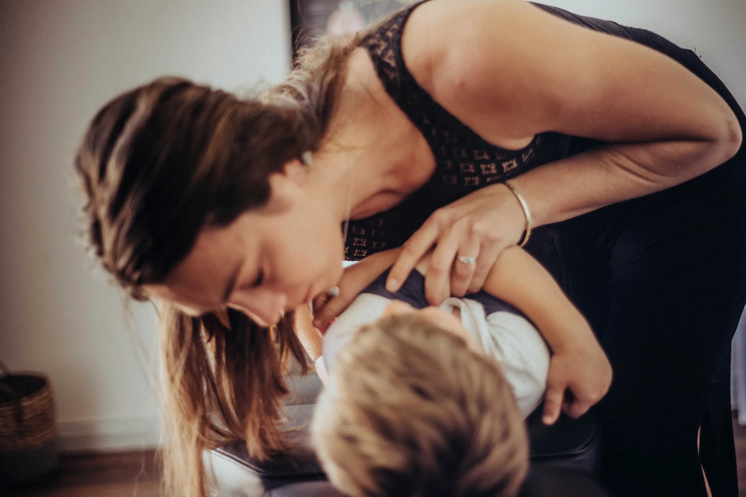 A woman with brown hair and glasses, wearing a black lace top, leaning over and kissing a young child's hand. The scene appears to be indoors with a neutral background.