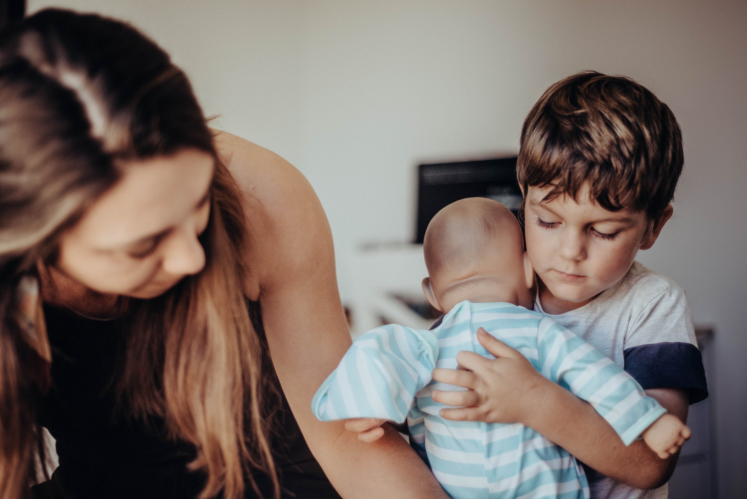 A woman and two young children are sharing a close, affectionate moment indoors. The woman is leaning over with long brown hair, and the children are hugging her and each other.