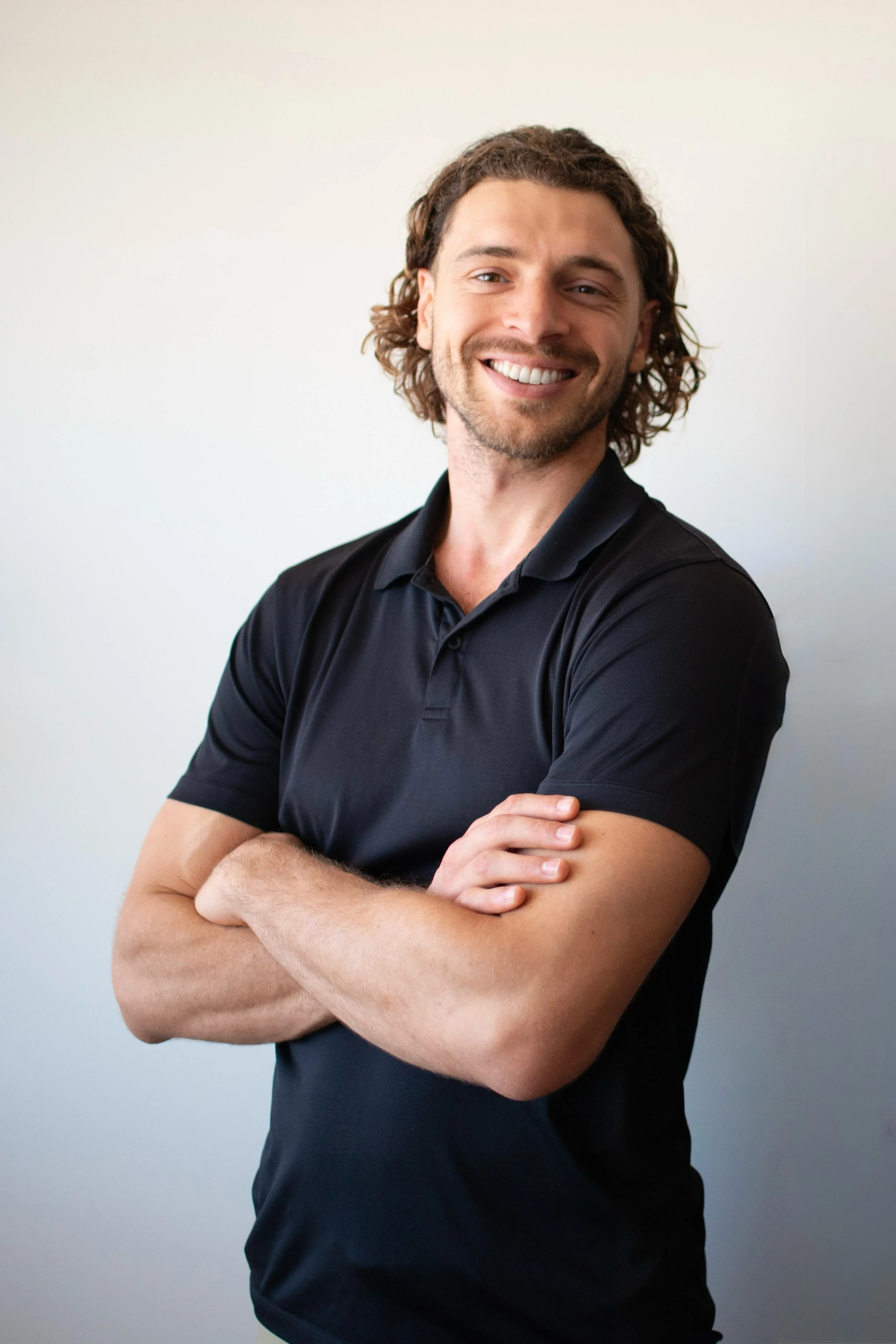 A smiling man with curly brown hair, crossed arms, wearing a black polo shirt, standing against a plain light-colored wall.