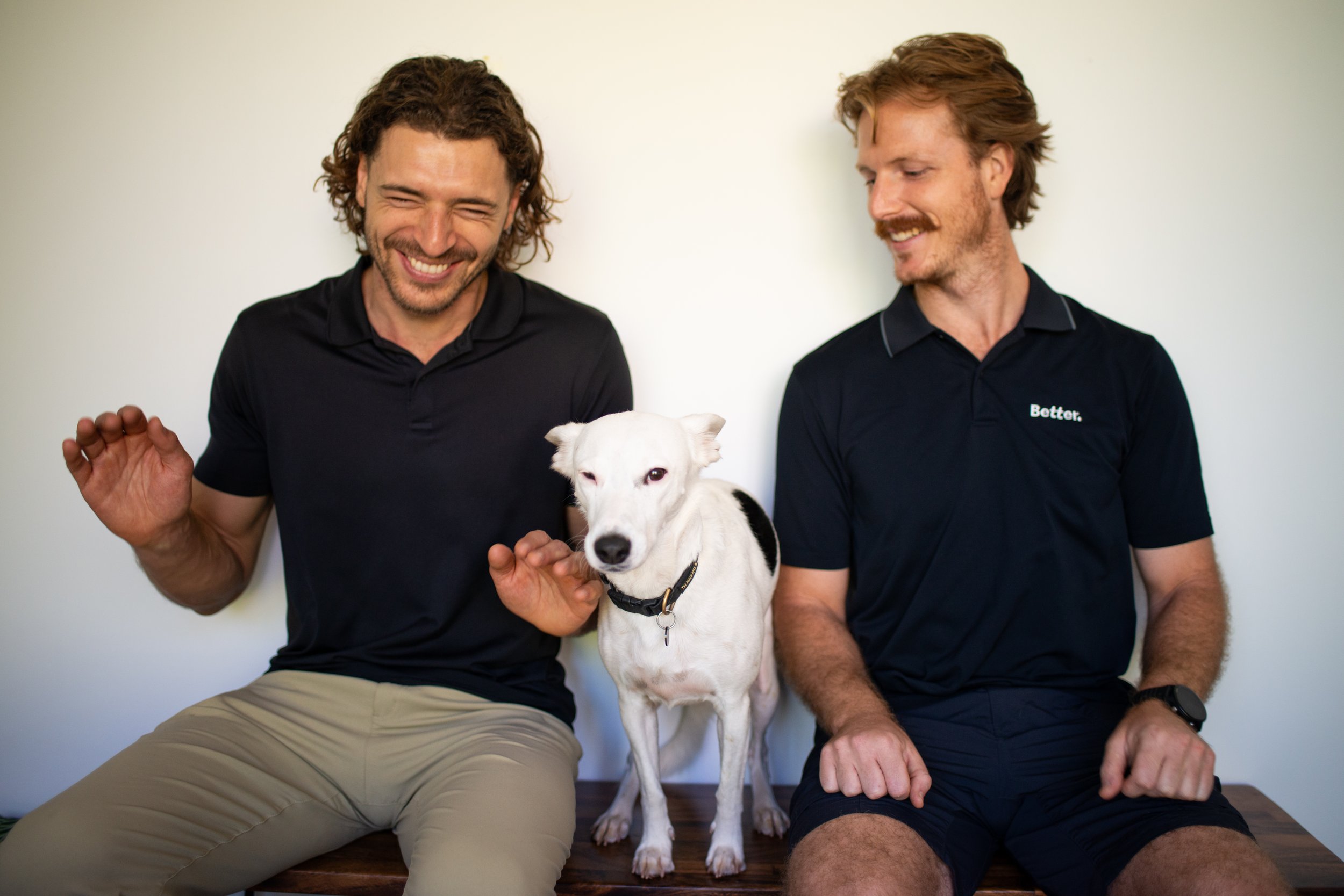 Two smiling men sitting on a wooden floor with a white dog between them, against a plain light-colored wall.