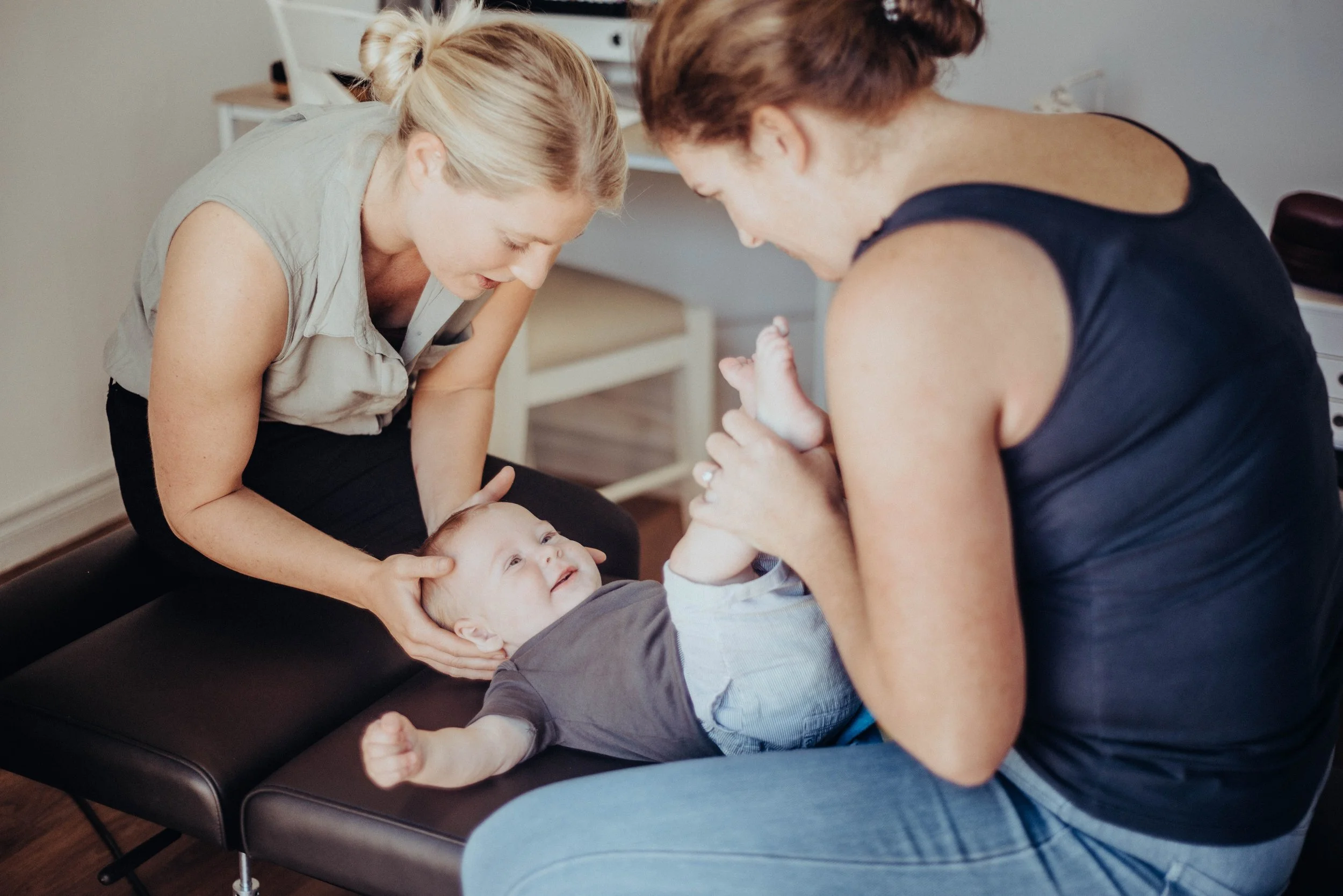 Two women are playing with and laughing at a young child lying on an examination table in a medical or dental office.