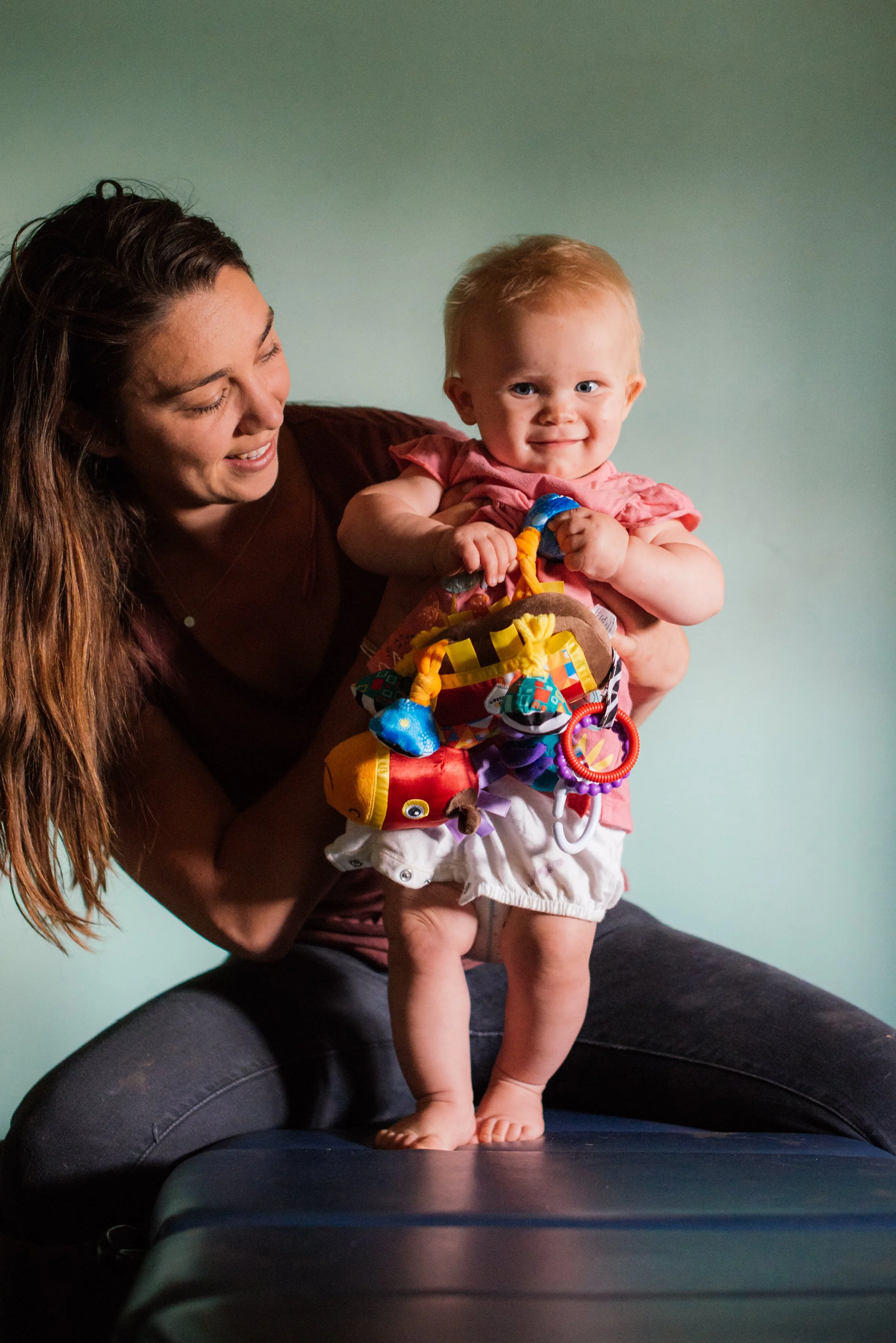 A woman holding a happy toddler girl with red hair, colorful toys, and white shorts. The woman is smiling, seated on a dark surface with a plain green wall in the background.