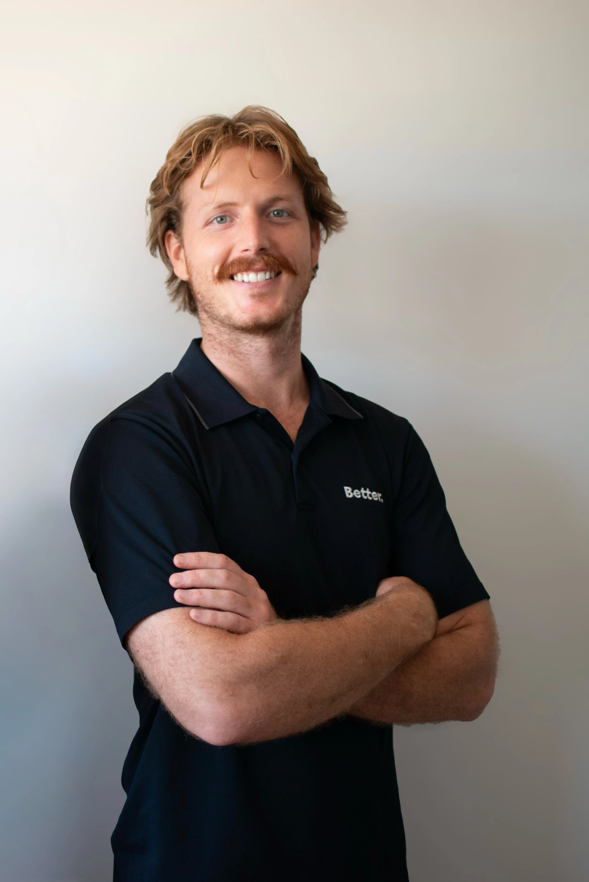 A man with red hair and a beard smiling, wearing a black polo shirt with white text, standing with arms crossed against a plain white wall.
