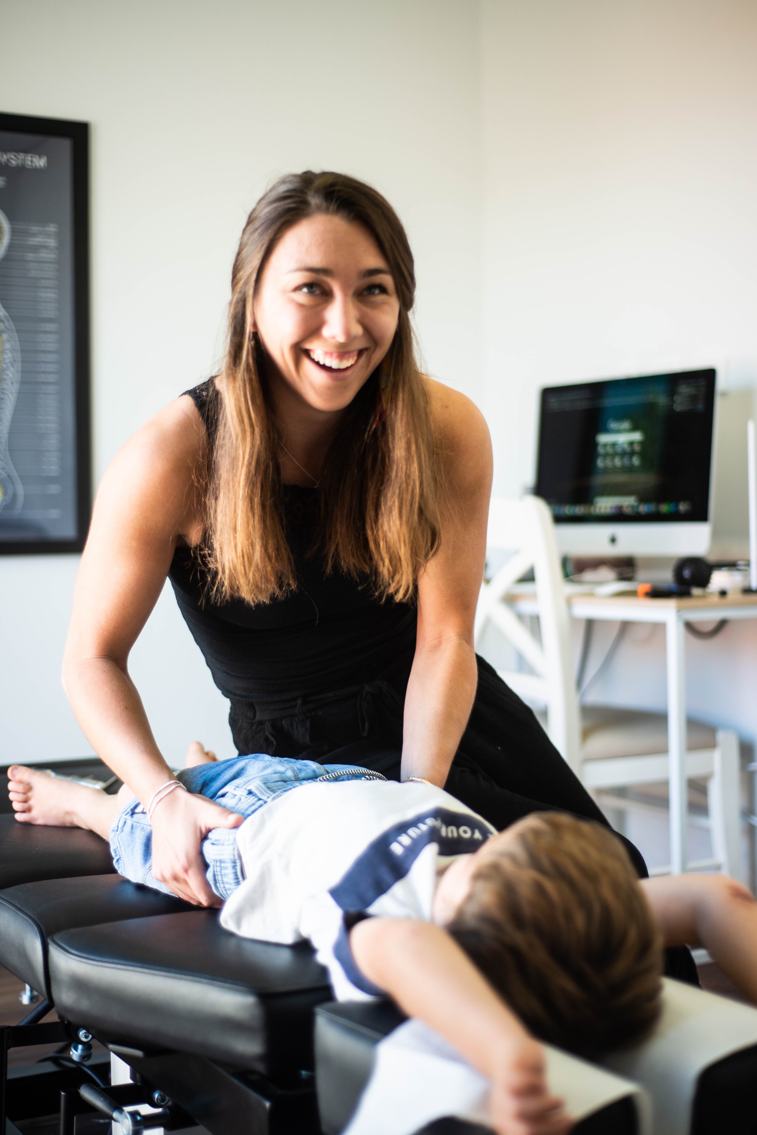 A woman with brown hair smiling, leaning over a child lying face down on a massage table, a computer in the background.