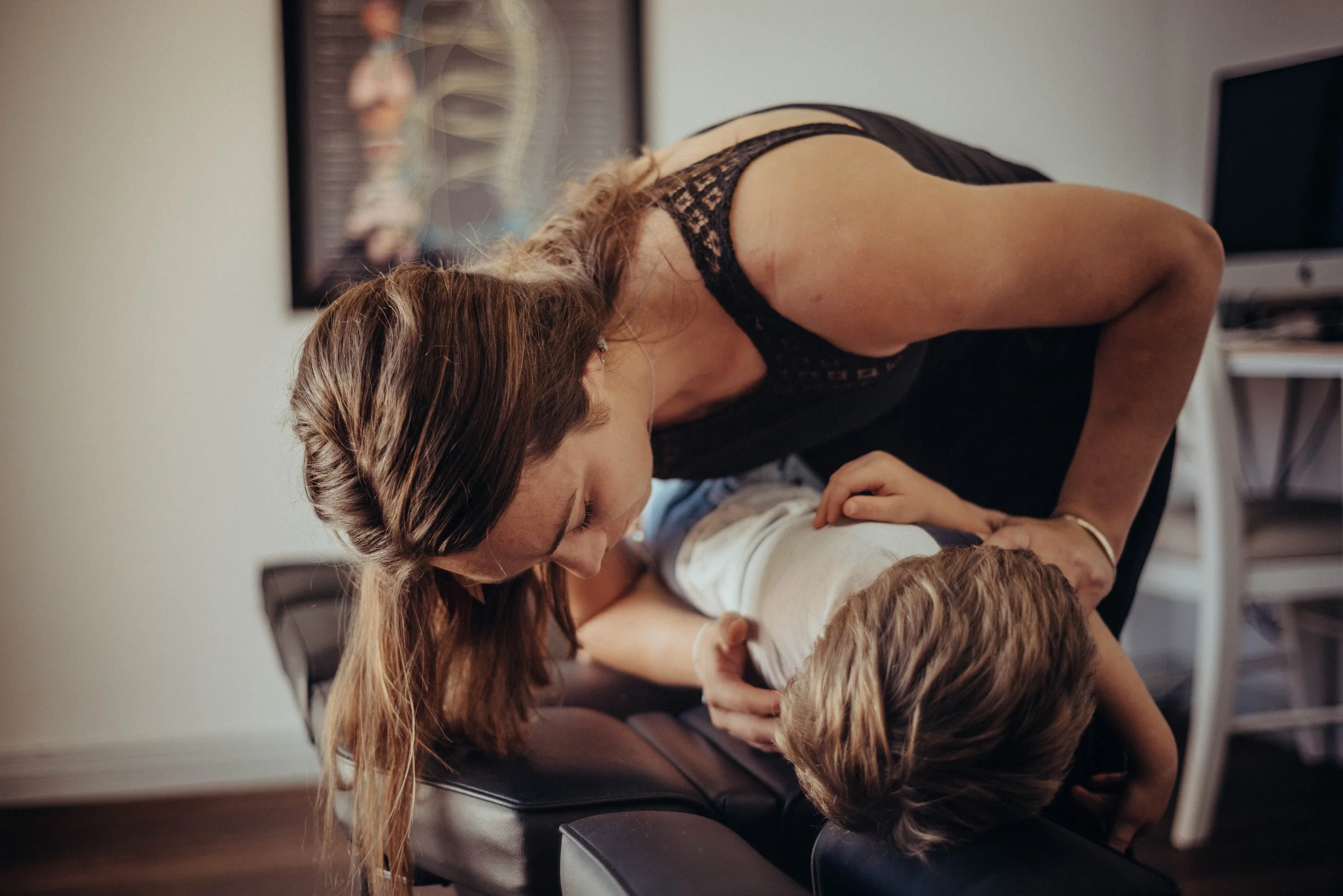 A woman with long hair leaning over a young child, engaging in a physical or playful activity in a room with medical diagrams on the wall, and a computer in the background.