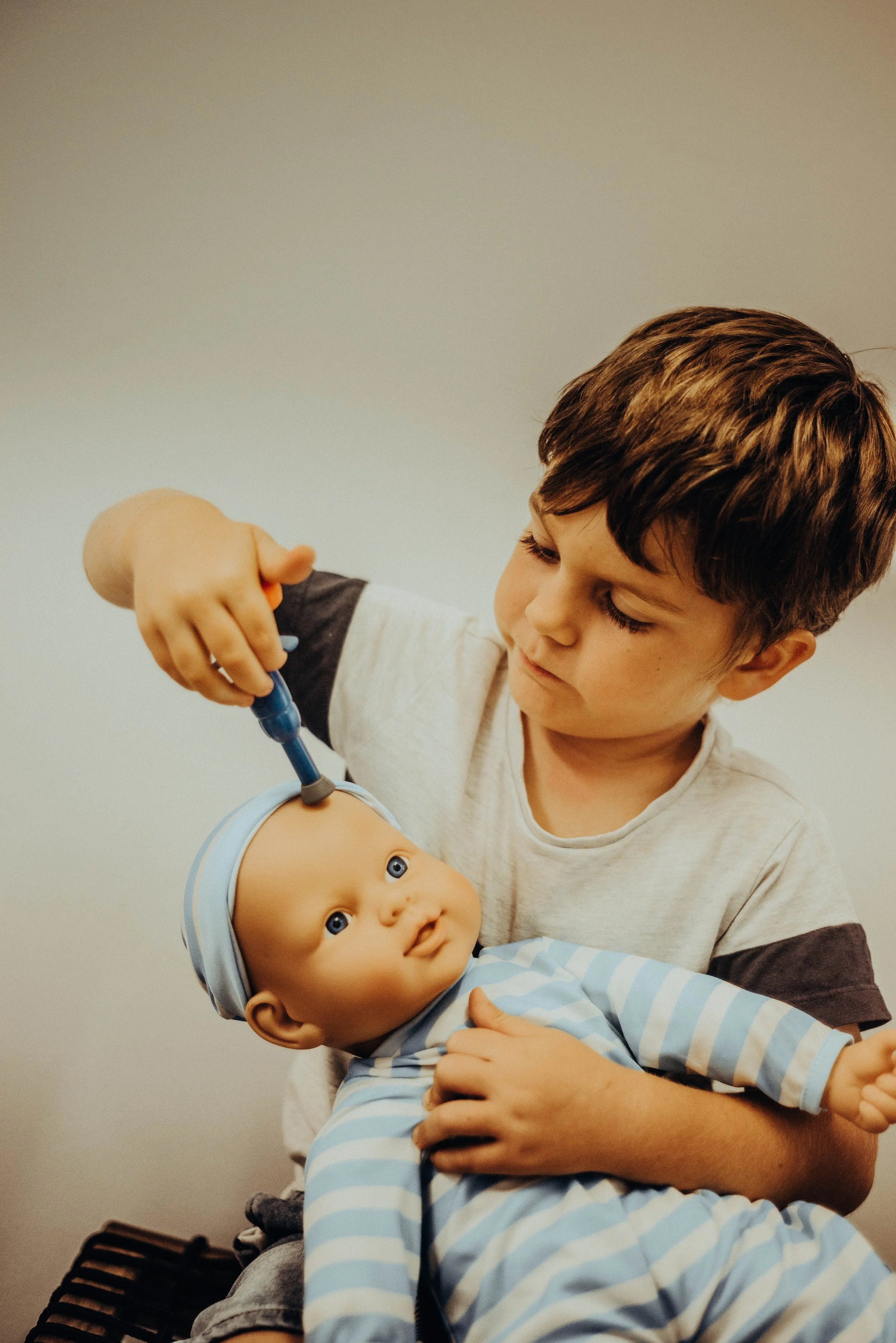 A young boy with brown hair is playing doctor with a medical doll, using a stethoscope, while sitting on a bench against a plain wall.