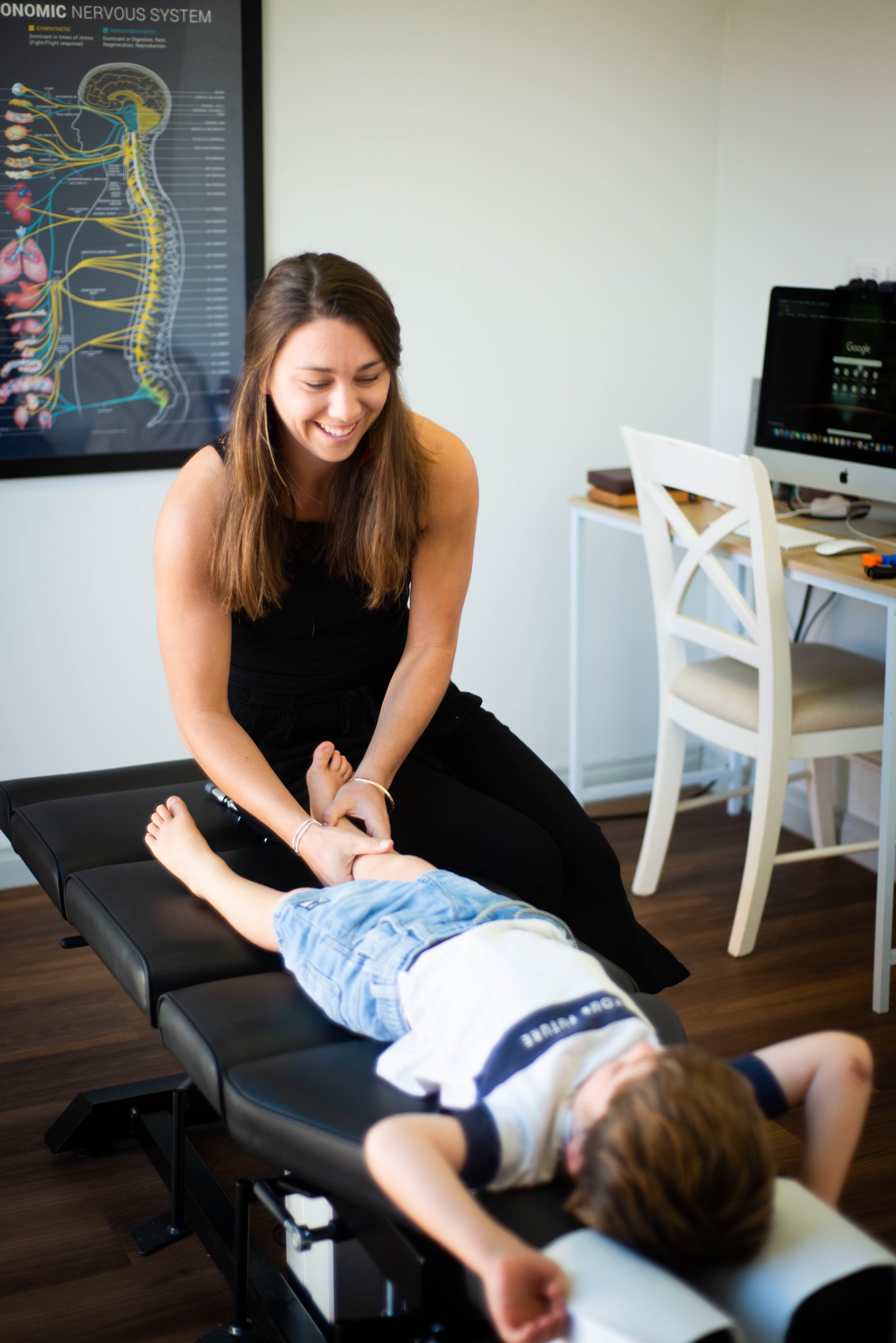 A woman with long brown hair smiling while holding a child's hand during a therapy session. A child is lying face down on a black therapy table, wearing a white shirt and blue jeans. A poster depicting the human nervous system is on the wall, and a computer and chair are on a small desk in the background.