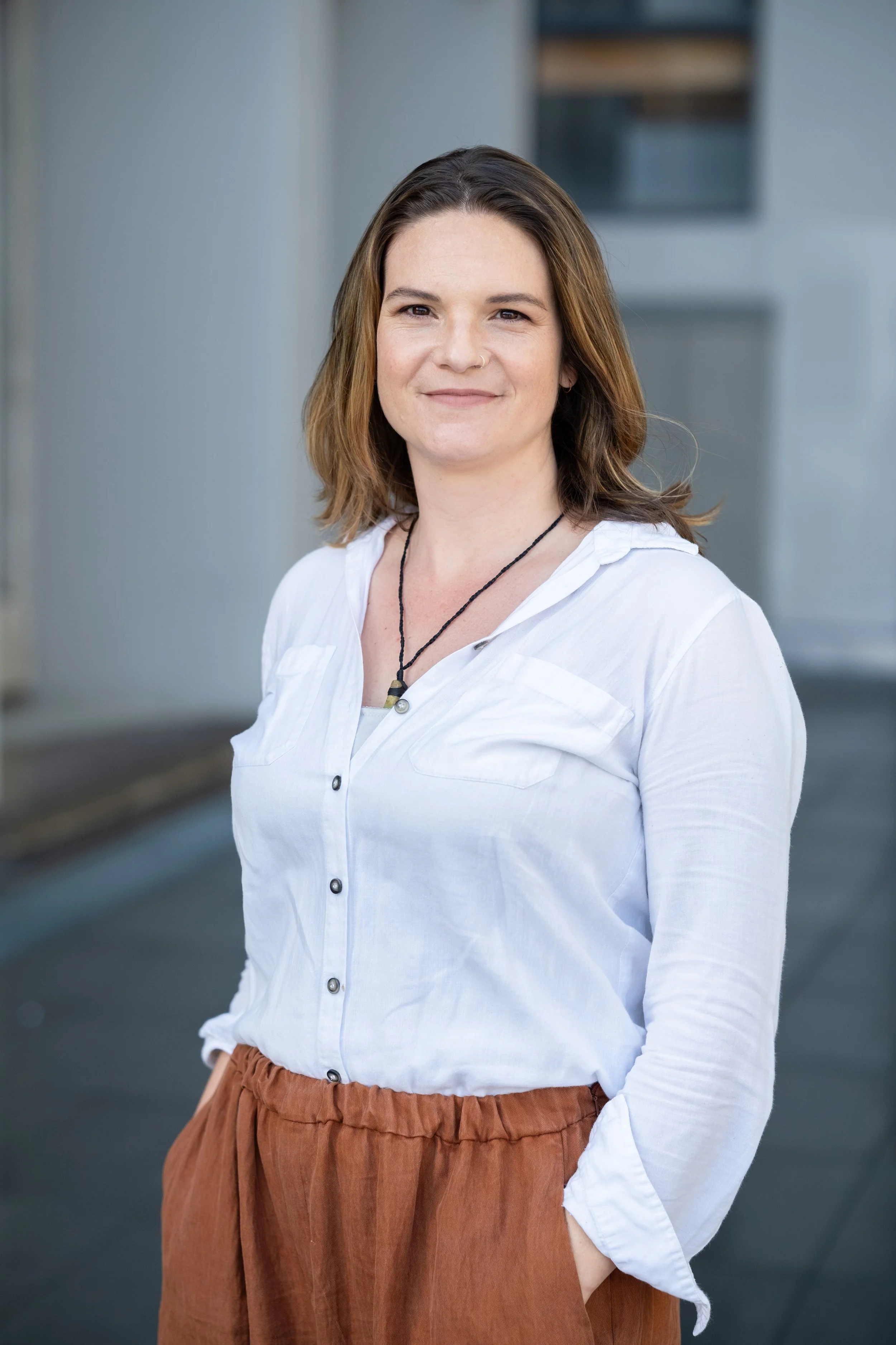 A woman with shoulder-length brown hair, wearing a white button-up shirt and rust-colored pants, stands outdoors with a blurred urban background.