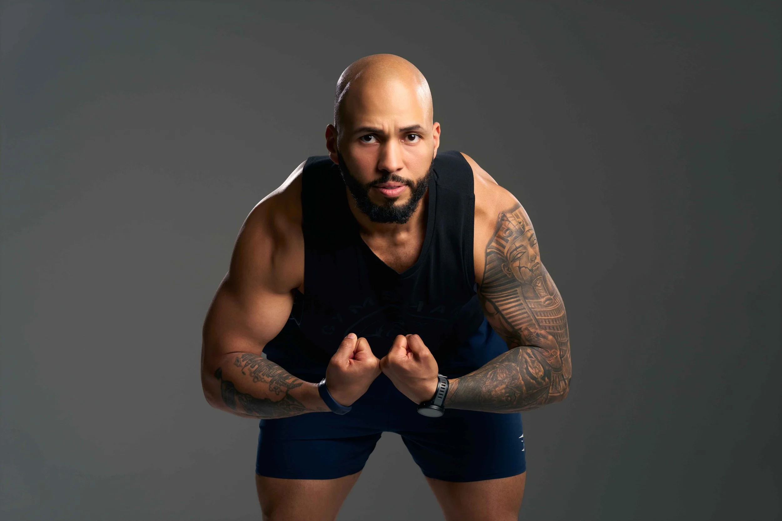 A muscular man with a beard and tattoos, wearing a black sleeveless shirt and shorts, is leaning forward with his hands clasped and looking at the camera against a gray background.