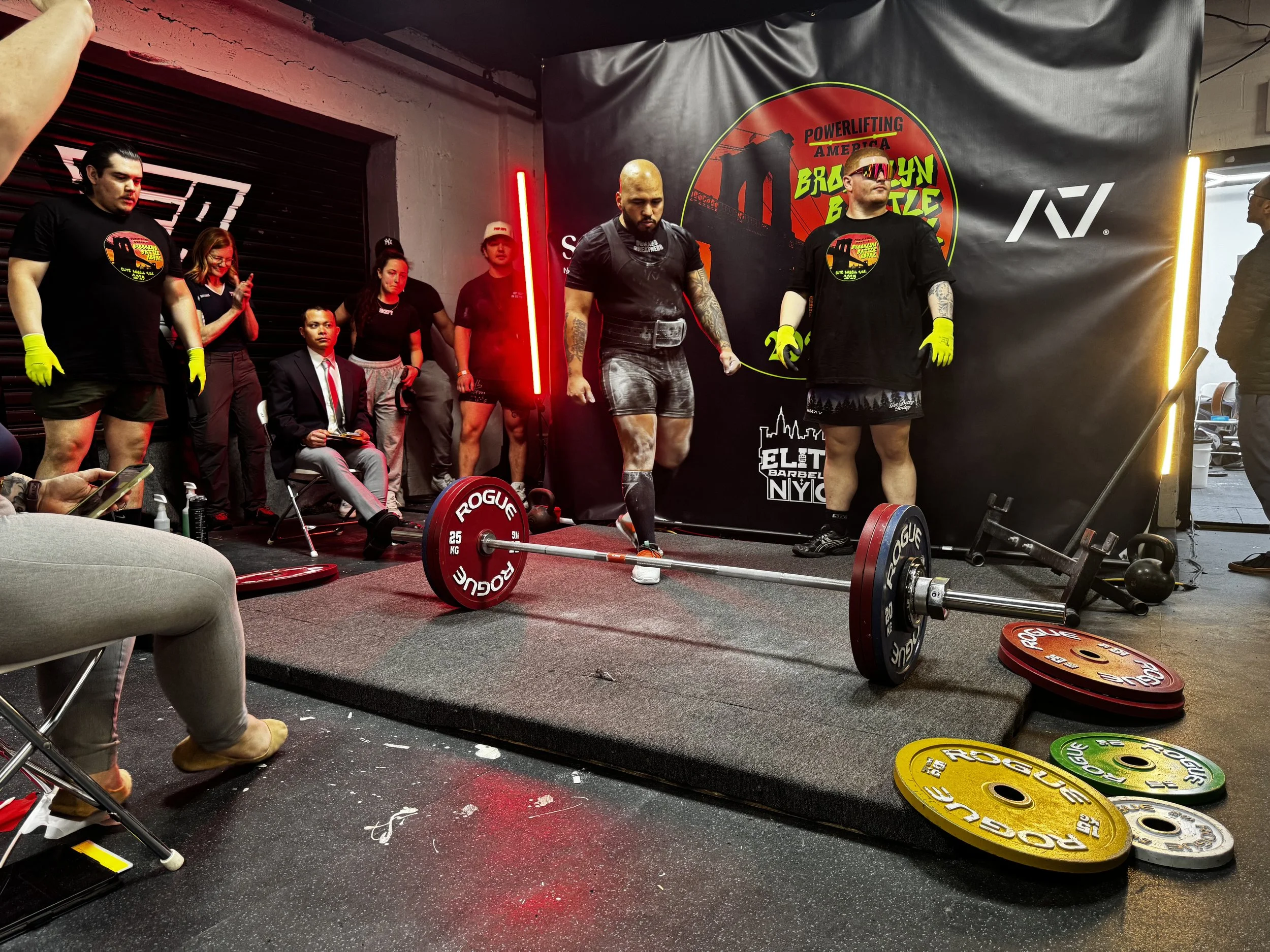 Group of people at a weightlifting competition with a black banner in the background advertising Powerlifting America Brazil Nut Battle. Two men in the foreground are preparing to lift weights, one with a barbell on the floor and another with glasses and tattoos, wearing black shirts and gloves. Several spectators and officials are standing or sitting nearby, some taking photos or clapping. Colorful weight plates are scattered on the floor.