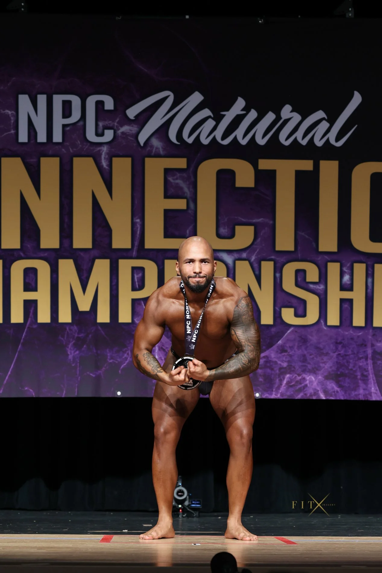 A male bodybuilder on stage at the NPC Natural Bodybuilding Championships, posing with a medal around his neck, in front of a purple background with event banner.