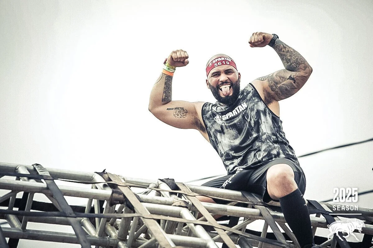 A man with tattoos wearing a headband and sports gear, flexing his muscles and sticking out his tongue while sitting on a metal obstacle at a Spartan race event in 2025.