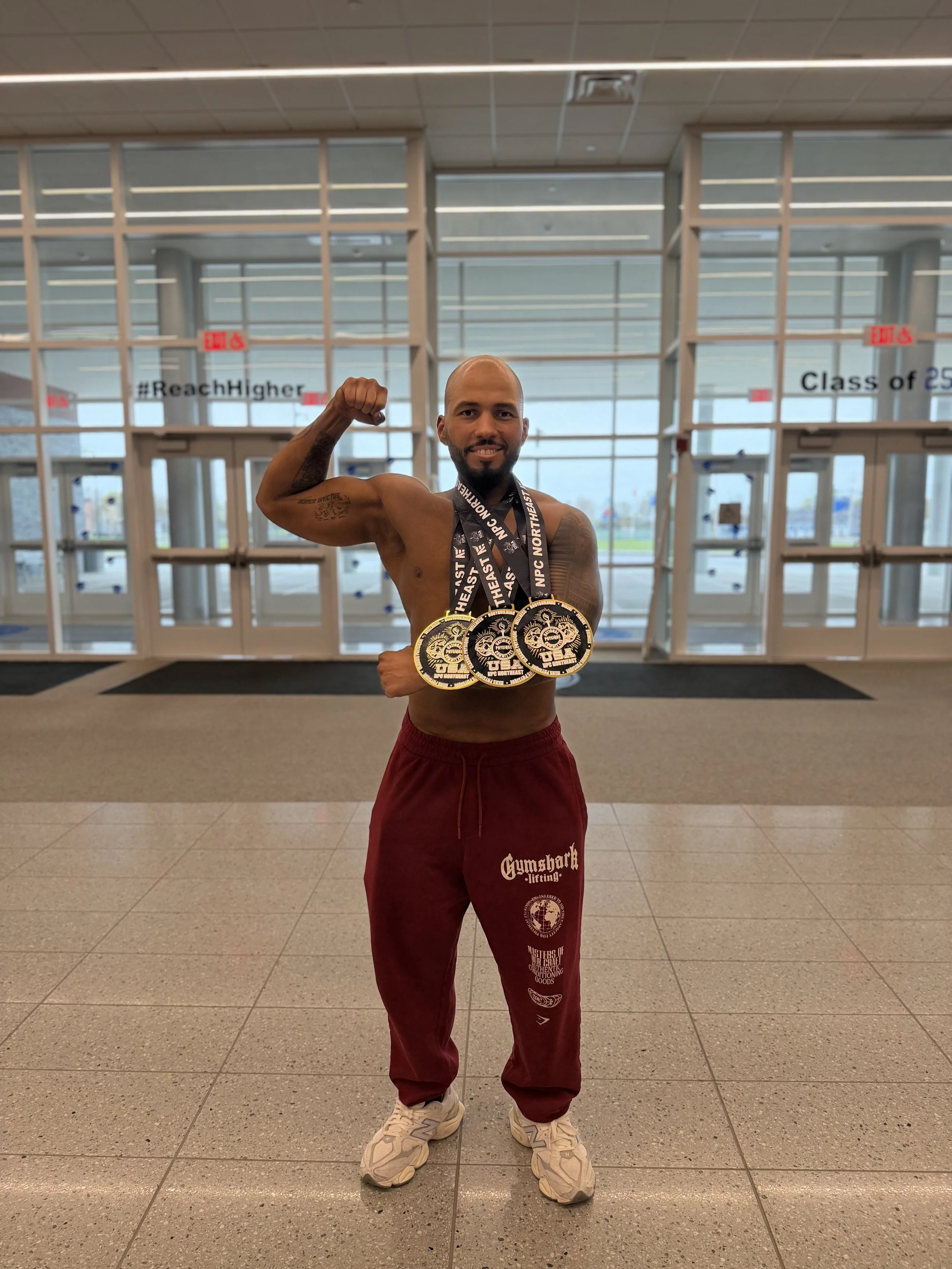 A muscular man with a beard and tattoos flexes his right arm, wearing medals around his neck, red sweatpants with gym logos, and sneakers, standing inside an airport terminal.