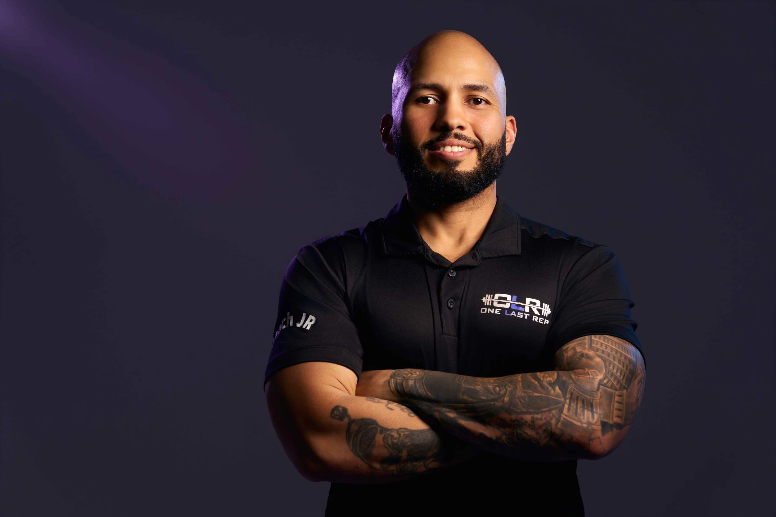 A smiling man with a bald head and a beard, wearing a black polo shirt with the logo 'One Last Rep' and crossed arms, standing against a dark background.