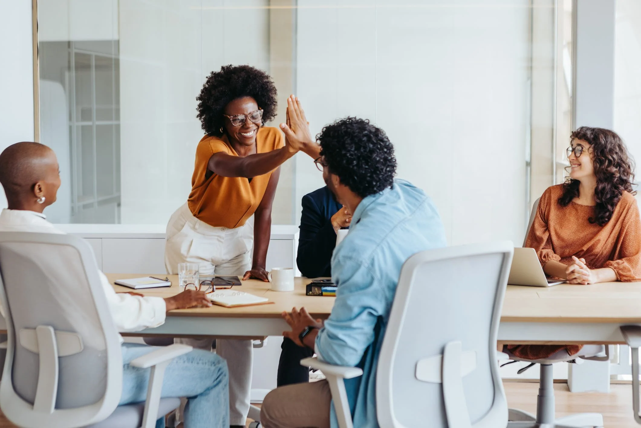 A diverse group of five women gathered around a conference table in a modern, bright office, engaging in a lively discussion. One woman is giving a high five to another who is smiling, while the others watch and listen, with notebooks, glasses, and a mug on the table.