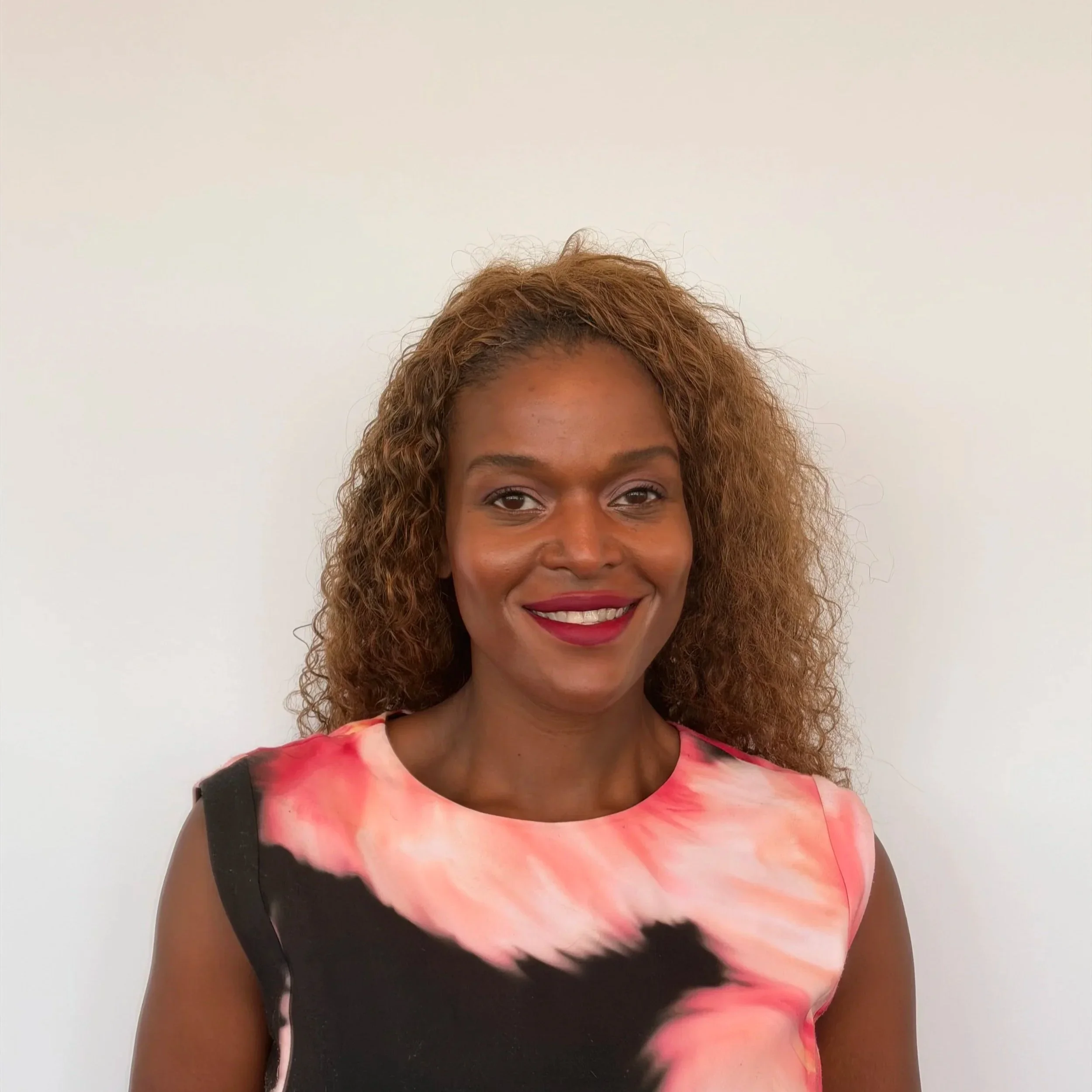 Professional headshot of a smiling woman with long curly hair wearing a pink and black patterned top.
