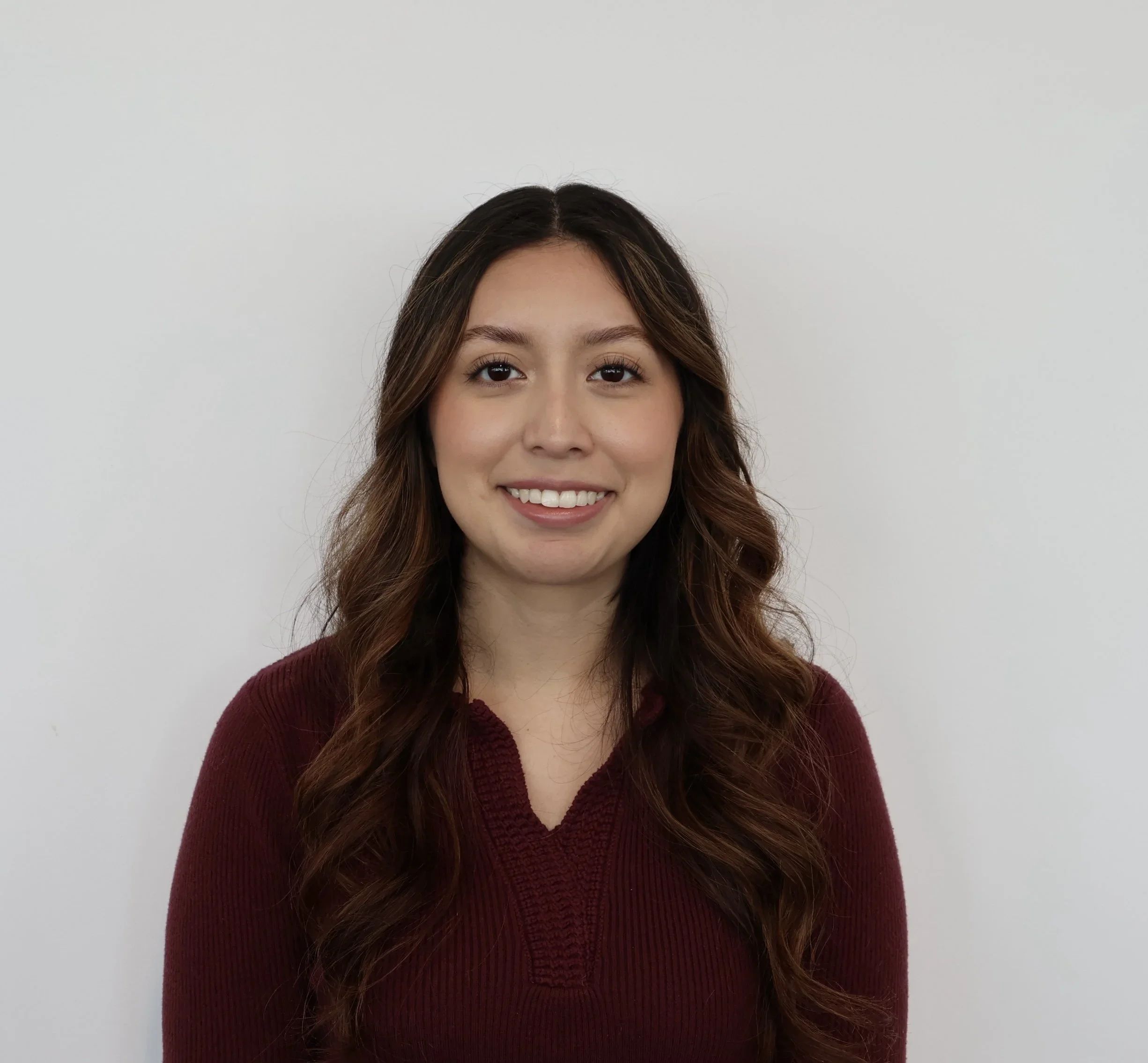 Professional headshot of a smiling woman with long wavy brown hair wearing a burgundy top.