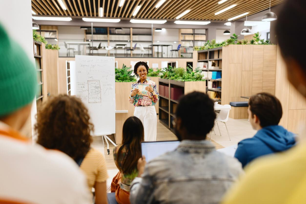 A woman giving a presentation or lecture to a group of diverse people in a modern, open office or classroom space with plants and bookshelves.