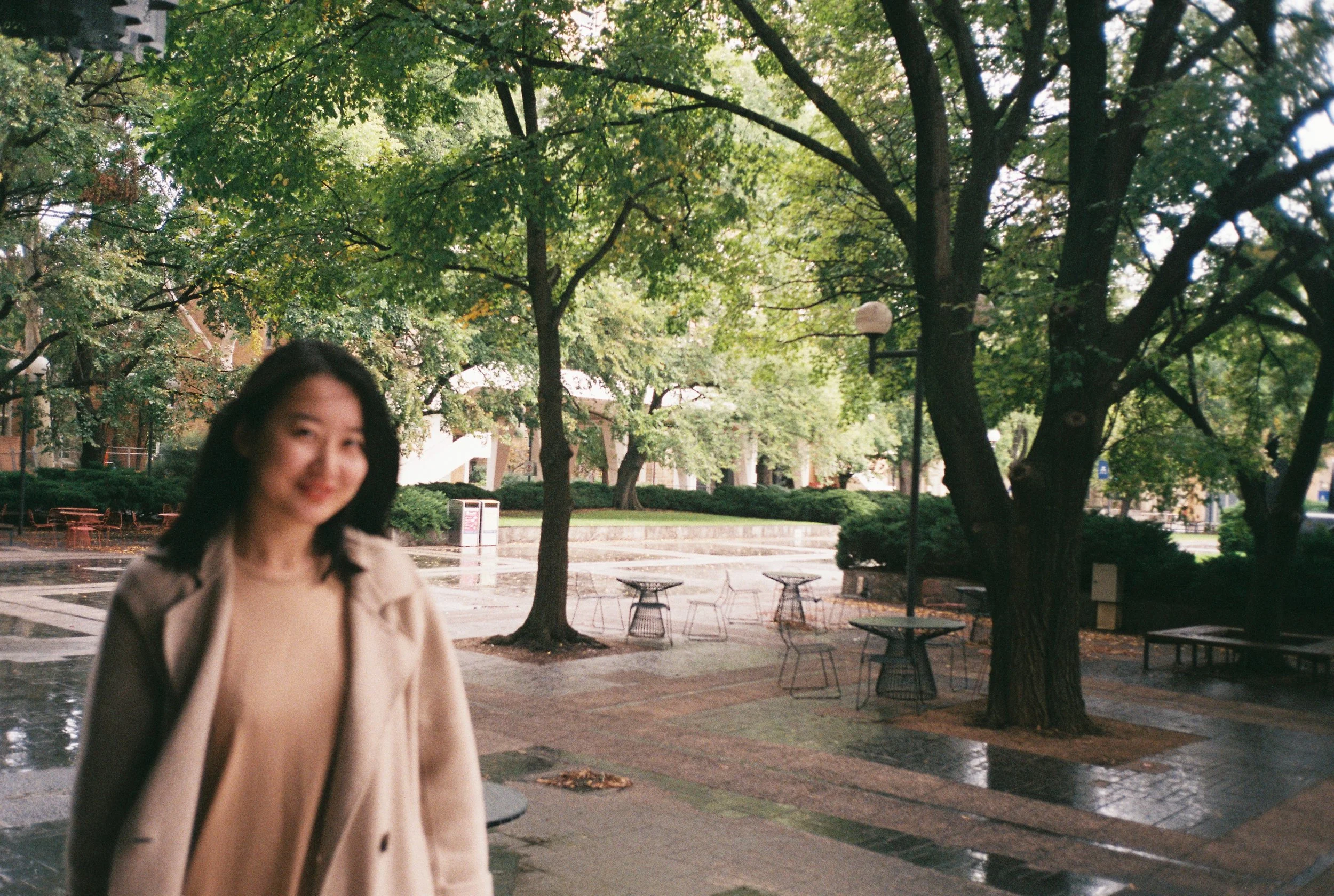 A woman with black hair smiling outdoors in a park with green trees and benches, on a rainy day.