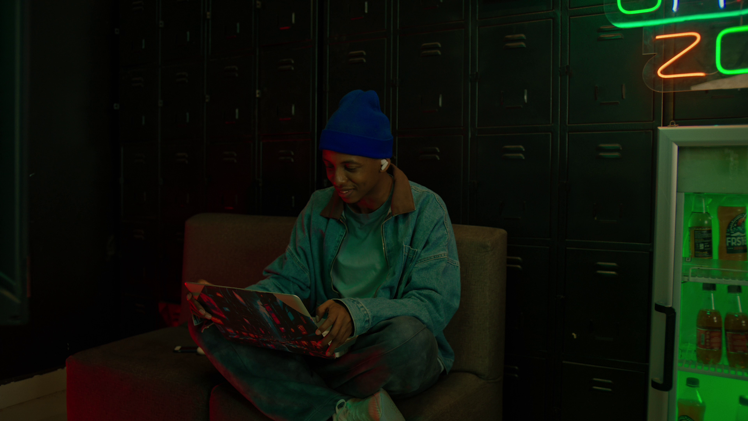 A young person with a blue beanie, denim jacket, and white earbuds sitting on a bench in a dimly lit room, looking at a tablet, with lockers and neon signage in the background.