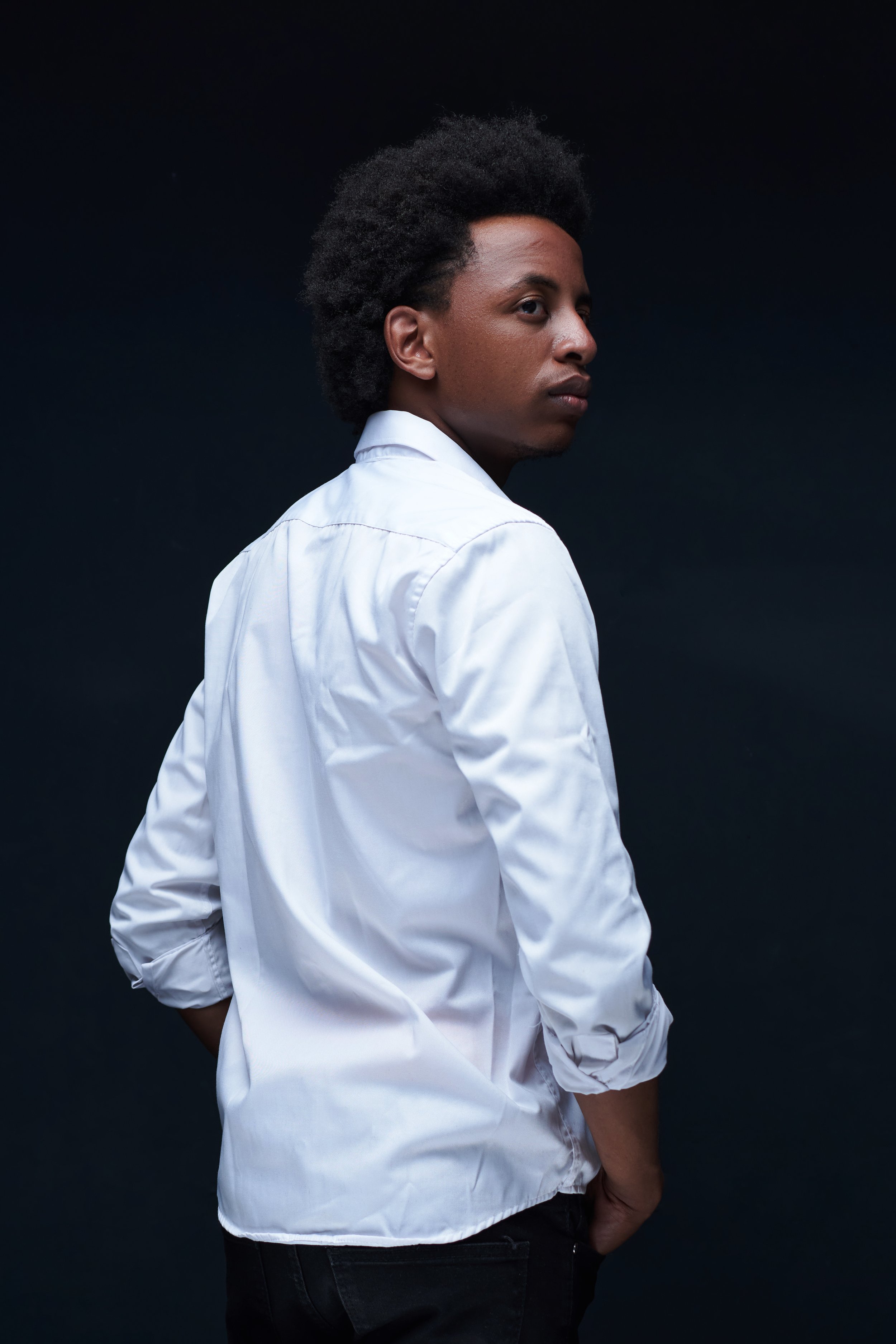 A young man with dark skin and curly black hair wearing a white shirt and dark pants standing in front of a dark background, looking over his shoulder.