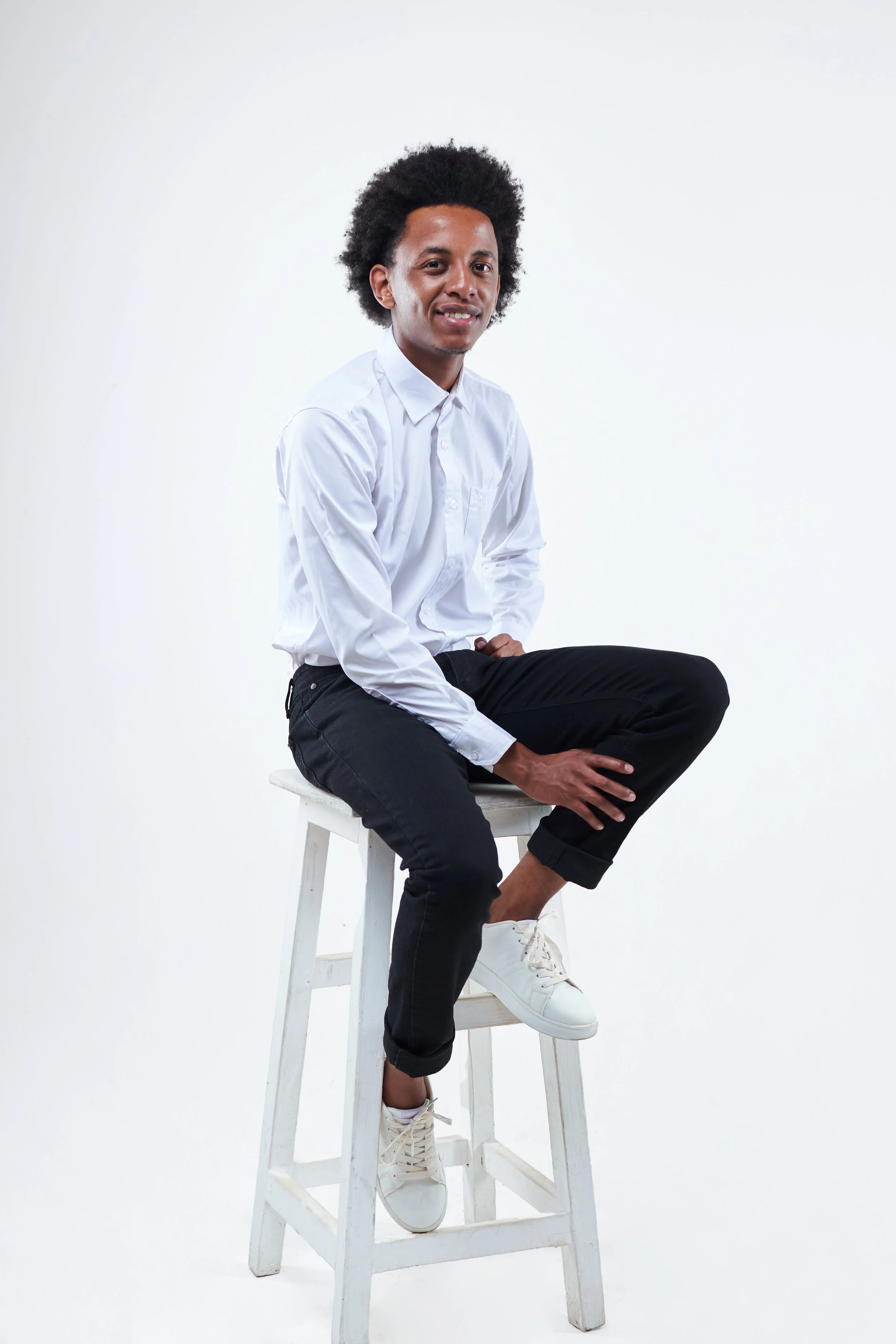 Young man with curly hair sitting on a white wooden stool, wearing a white shirt, black pants, and white sneakers, against a plain white background.