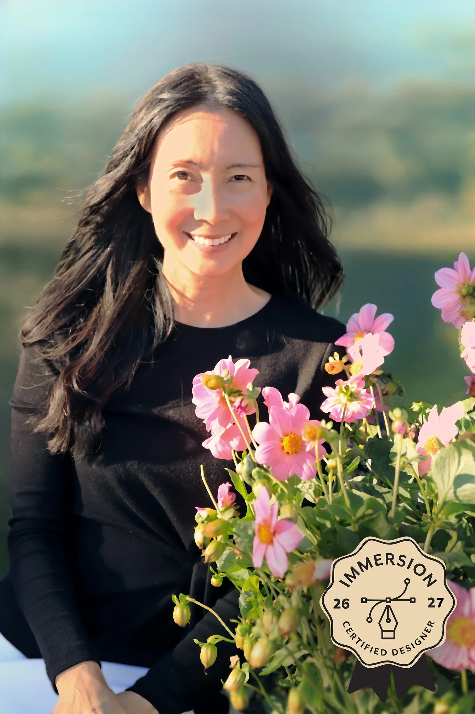 A woman with long dark hair smiling while holding a bouquet of pink flowers, standing outdoors with a blurred green background.