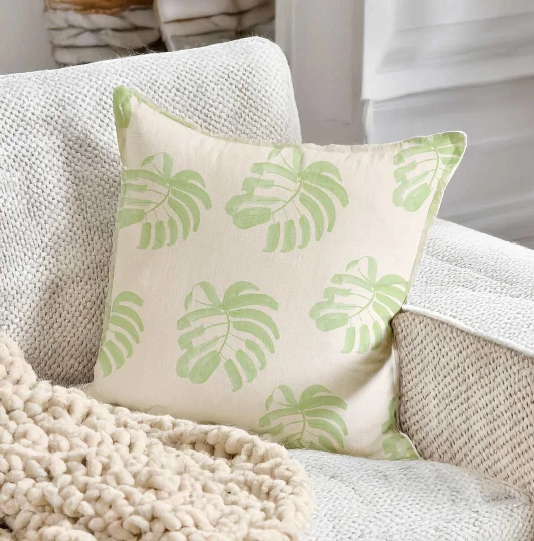 A decorative pillow with green Monstera leaf patterns on a white background, placed on a textured white couch.