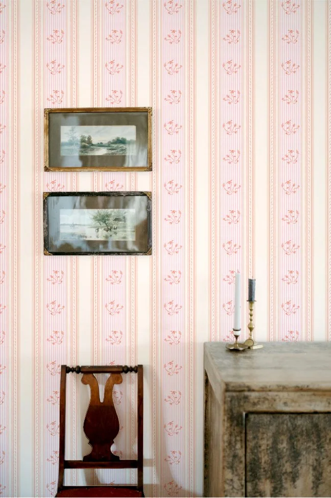 Vintage room with pink floral wallpaper, two landscape paintings framed in gold hang on the wall, an antique wooden chair with a carved backrest, and a weathered wooden cabinet with a brass candlestick and candle on top.