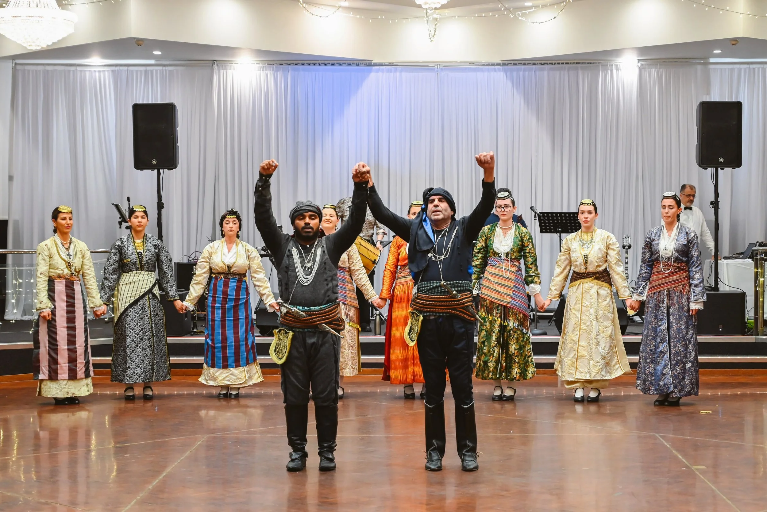 Performers in traditional clothing showing a cultural dance on stage with a white curtain backdrop and microphones, two large speakers, and musical instruments.