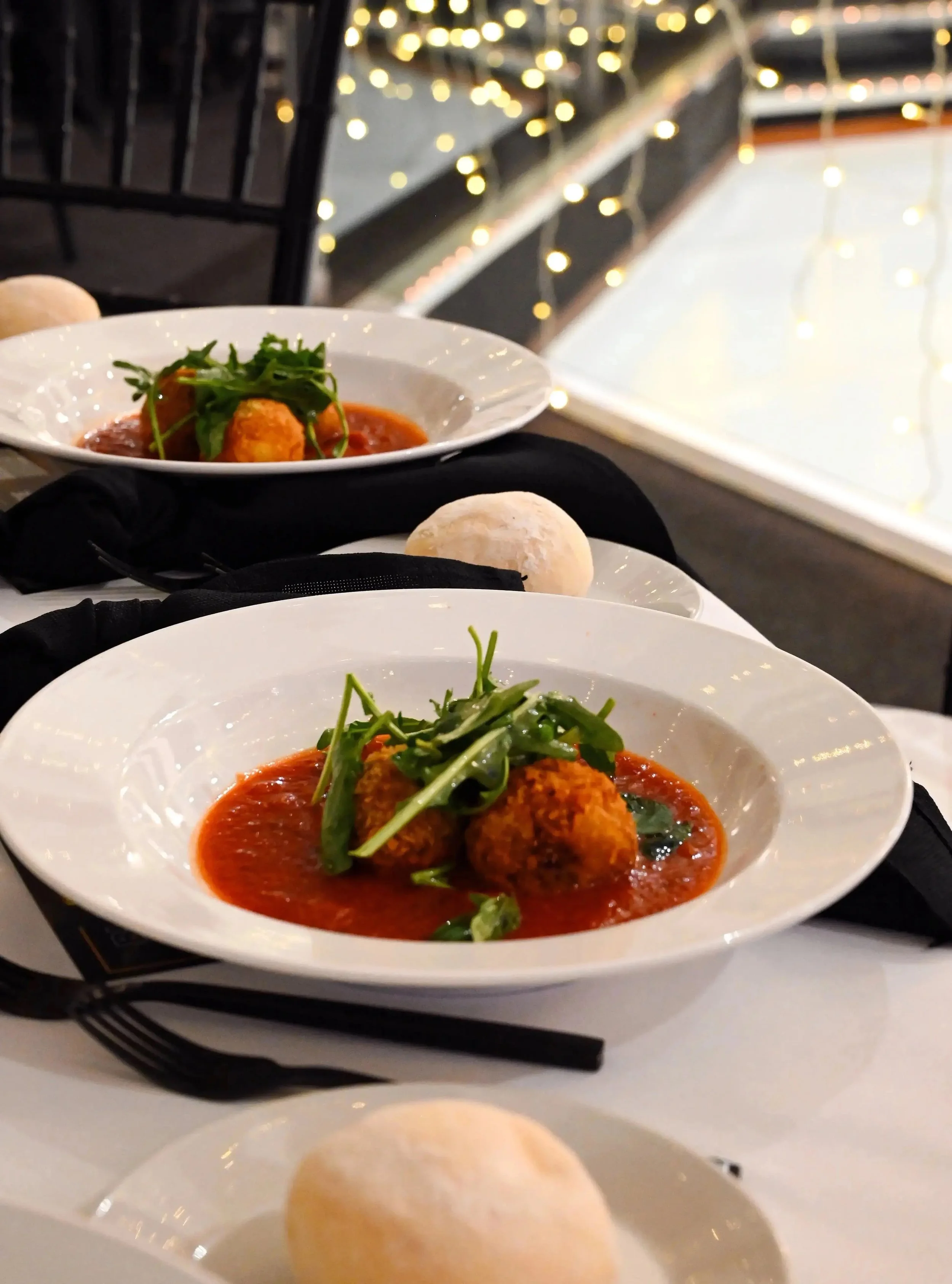 Dish of meatballs in marinara sauce garnished with arugula, served on a white plate, with a bread roll, in a dimly lit restaurant with fairy lights in the background.