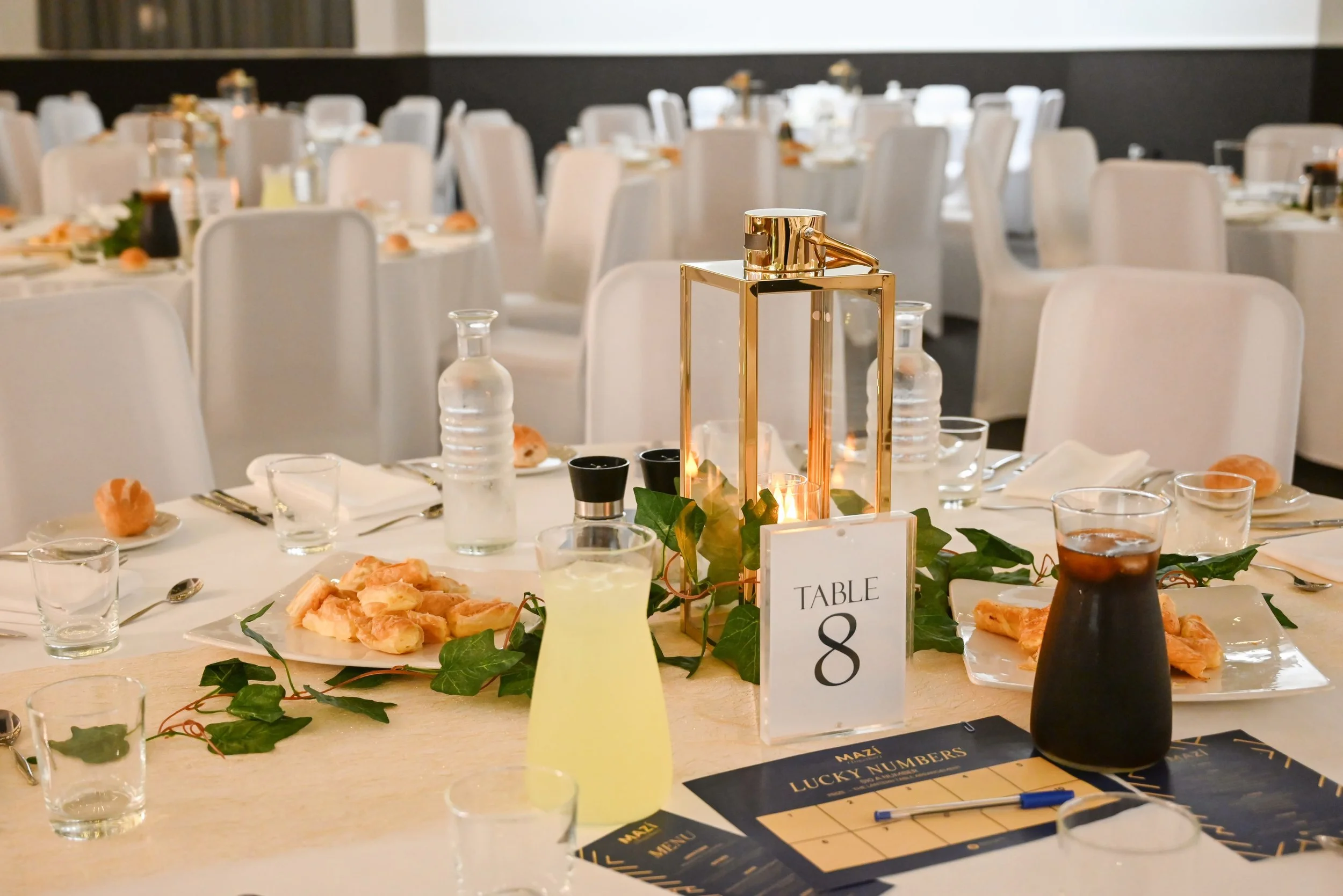 A banquet table decorated with a tall gold lantern surrounded by greenery, with plates of food, glasses, and utensils set for a meal, and a table number card reading 'Table 8' on the table.