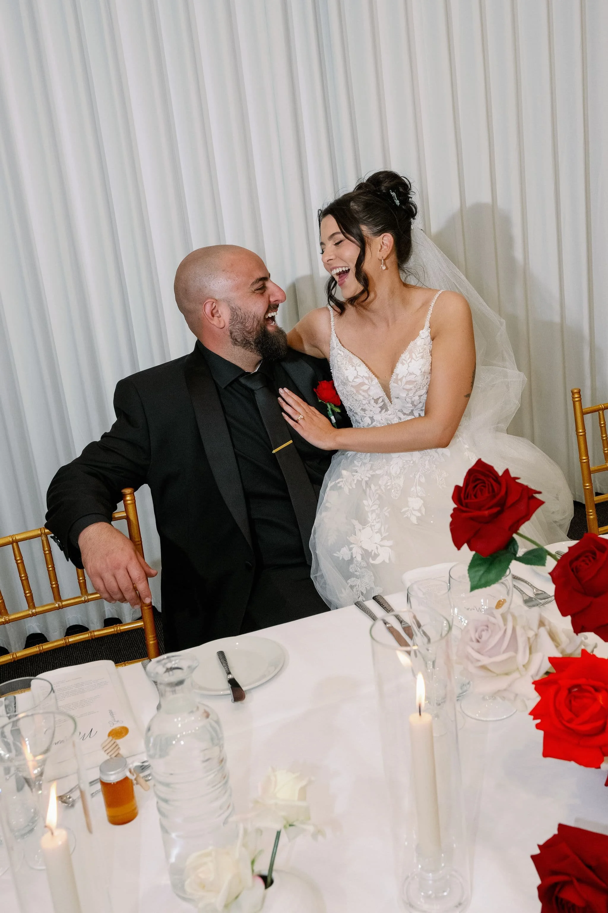 A bride and groom laughing together at their wedding reception, sitting at a table decorated with red and white flowers, candles, and glassware at Donato reception centre in Adelaide