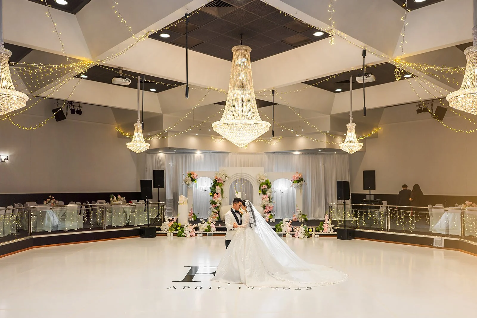 A bride and groom are dancing in the center of a wedding reception hall decorated with elegant chandeliers and fairy lights. The background features a floral arch and tables with white tablecloths on one side at Donato the venue in Adelaide
