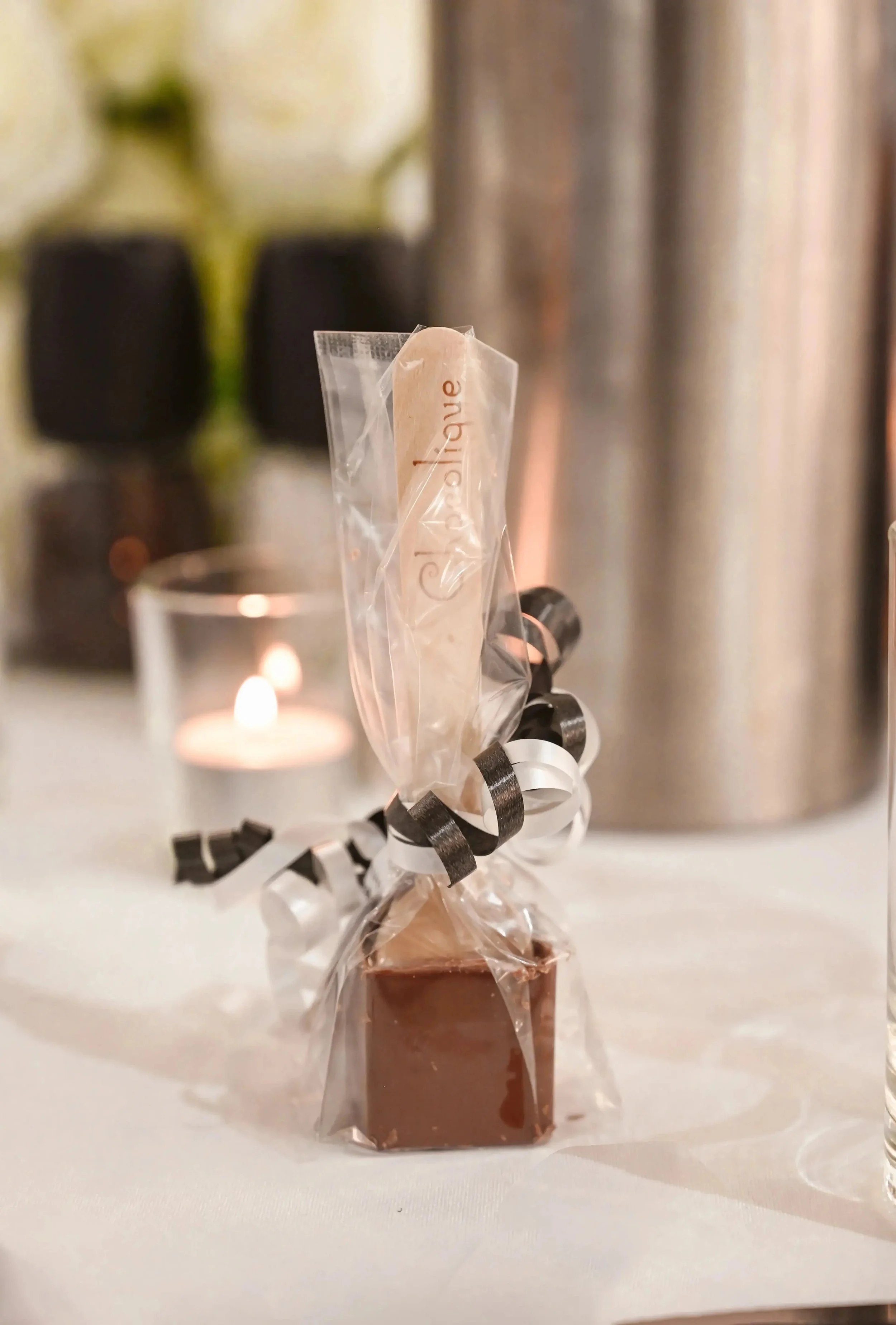 A square piece of chocolate cake wrapped in clear plastic and tied with black and white curled ribbon, with a beige dessert spoon in its wrapper. In the background, there are blurred plants and metallic objects, along with a lit candle.