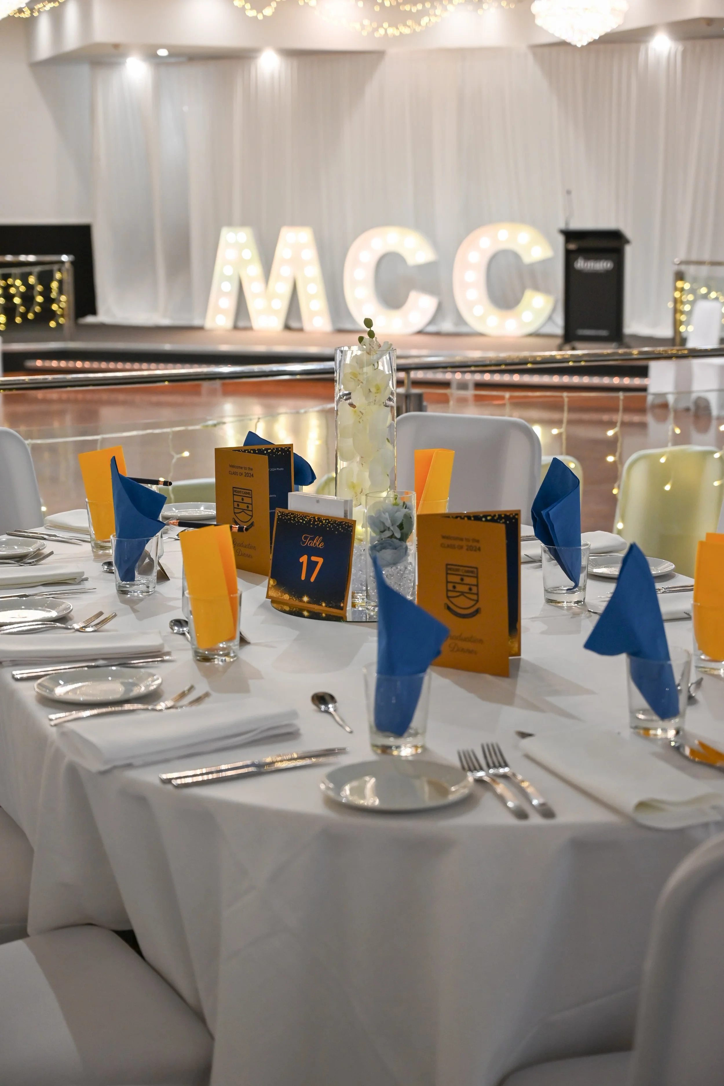 A banquet hall with decorated tables, including a centerpiece with white flowers, blue and yellow napkins, and place cards. In the background, large illuminated letters spell out 'MCC' on a stage with white drapery and string lights.