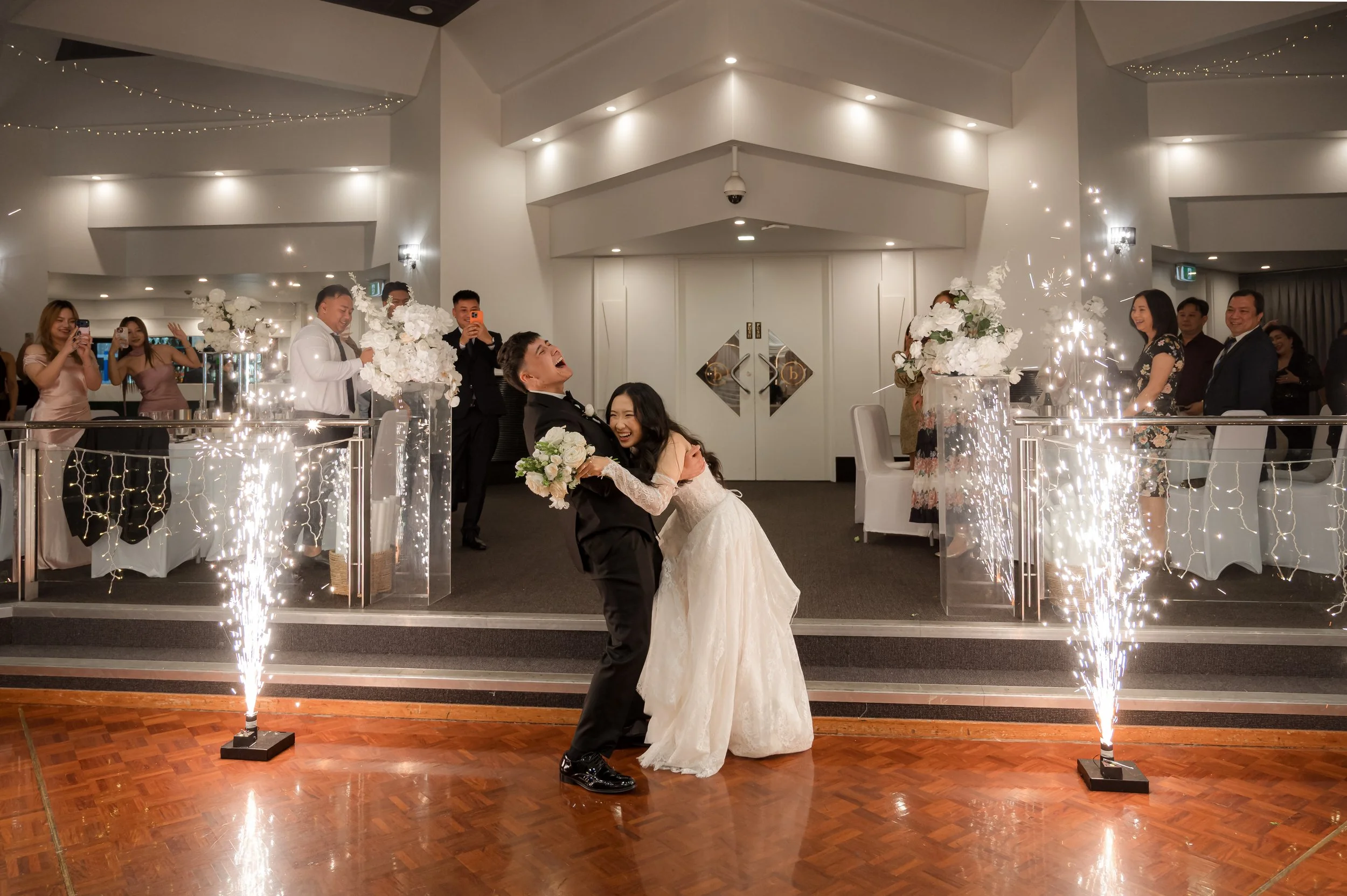 A newlywed couple dancing and hugging in front of a crowd at their wedding reception, with sparklers on the floor and fairy lights hanging from the ceiling.