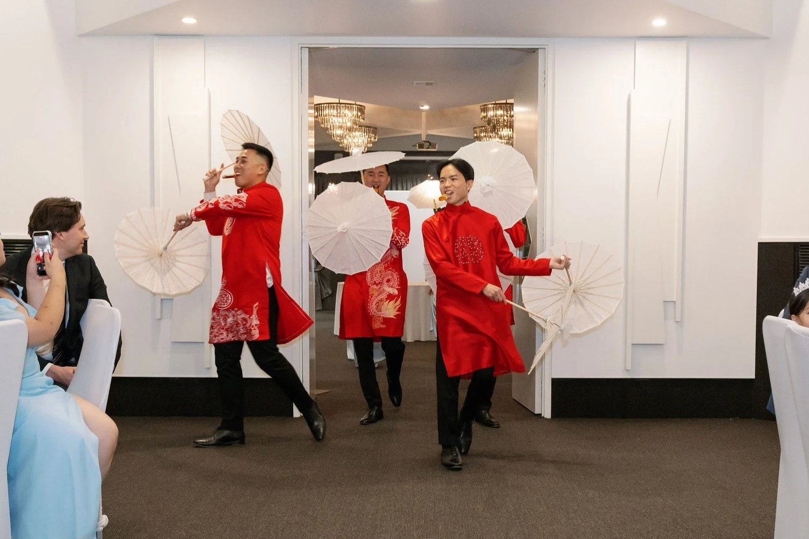 Three men dressed in red traditional Vietnamese áo dài are performing a dance with white umbrellas at a wedding reception, with guests sitting and watching in a decorated indoor venue.