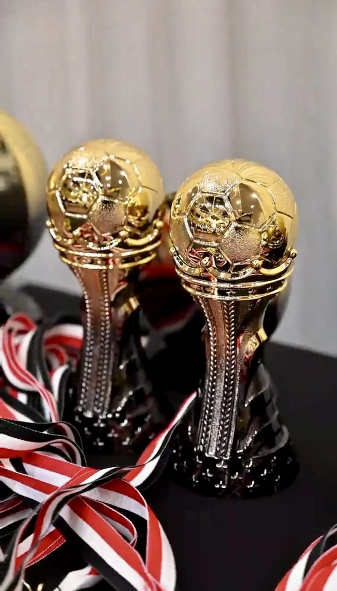 Close-up of two gold-colored trophies with honeycomb patterns on top, placed on a black surface with red, white, and black ribbons around them.