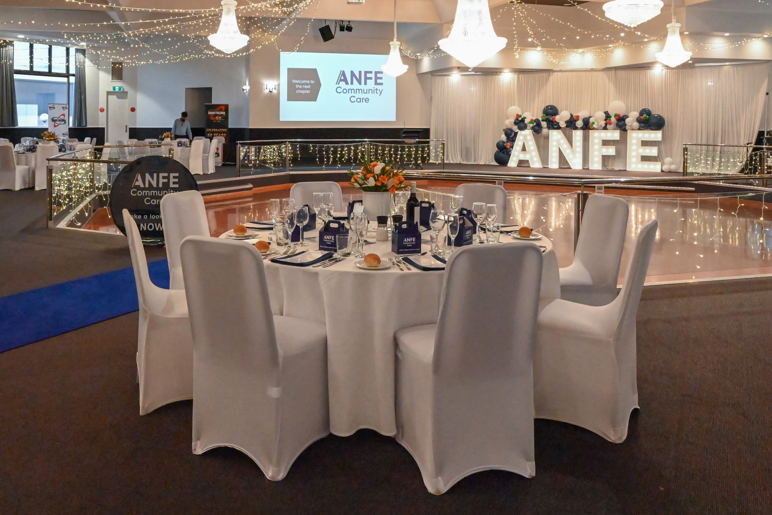 Banquet hall decorated for an event, featuring a stage with balloons and illuminated letters spelling 'ANFE,' round tables with white tablecloths, chairs, glasses, and centerpieces, string lights hanging from the ceiling, a large screen displaying th