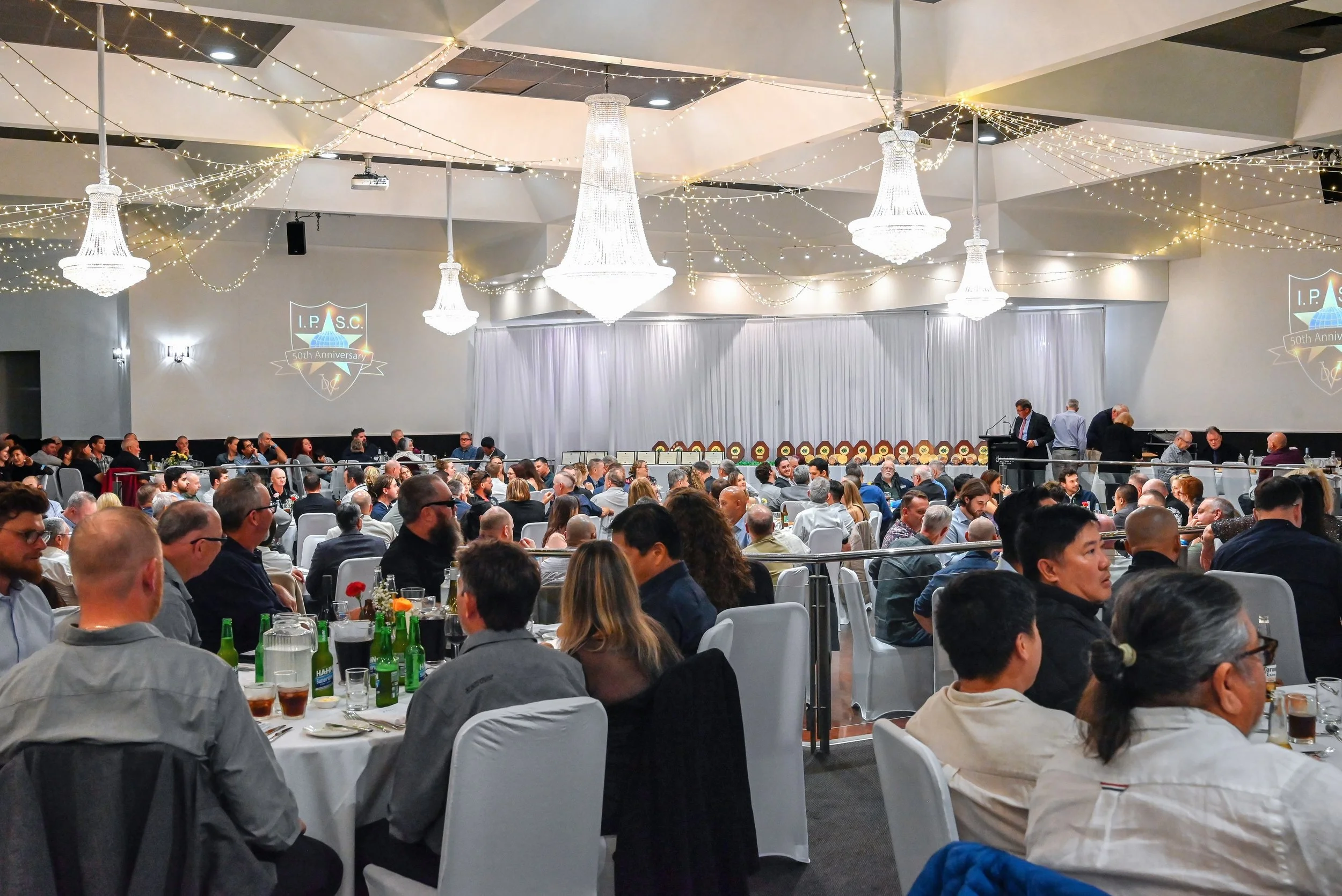 A large banquet hall filled with numerous round tables occupied by people attending a formal event. The ceiling is decorated with string lights and large chandeliers. There is a stage with a white curtain backdrop, and a projector is displaying the l