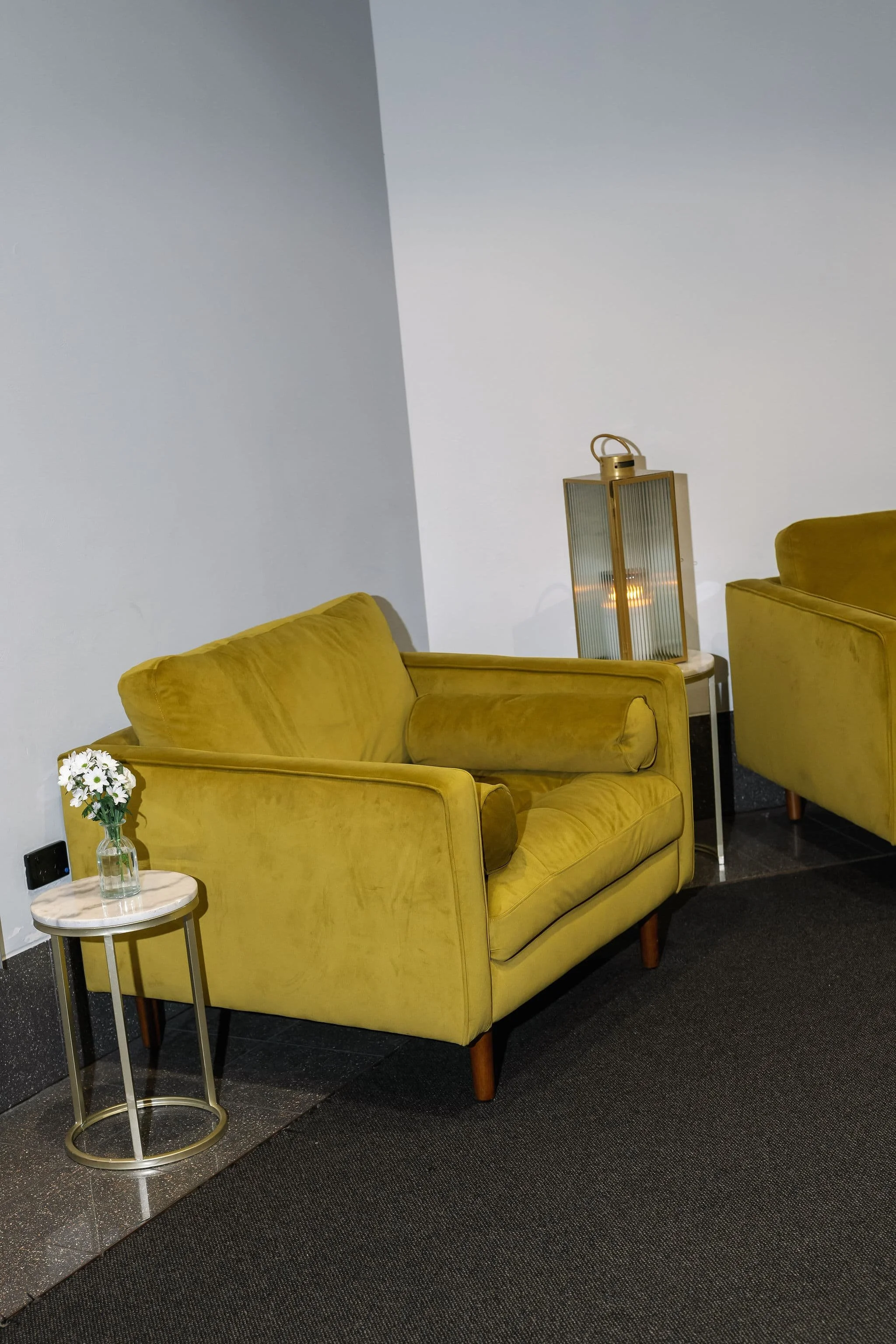 A room with two yellow velvet armchairs and a small side table with a glass vase of white flowers. A tall, gold-framed glass lantern with a candle inside is positioned between the chairs, against a plain white wall with a dark carpeted floor.