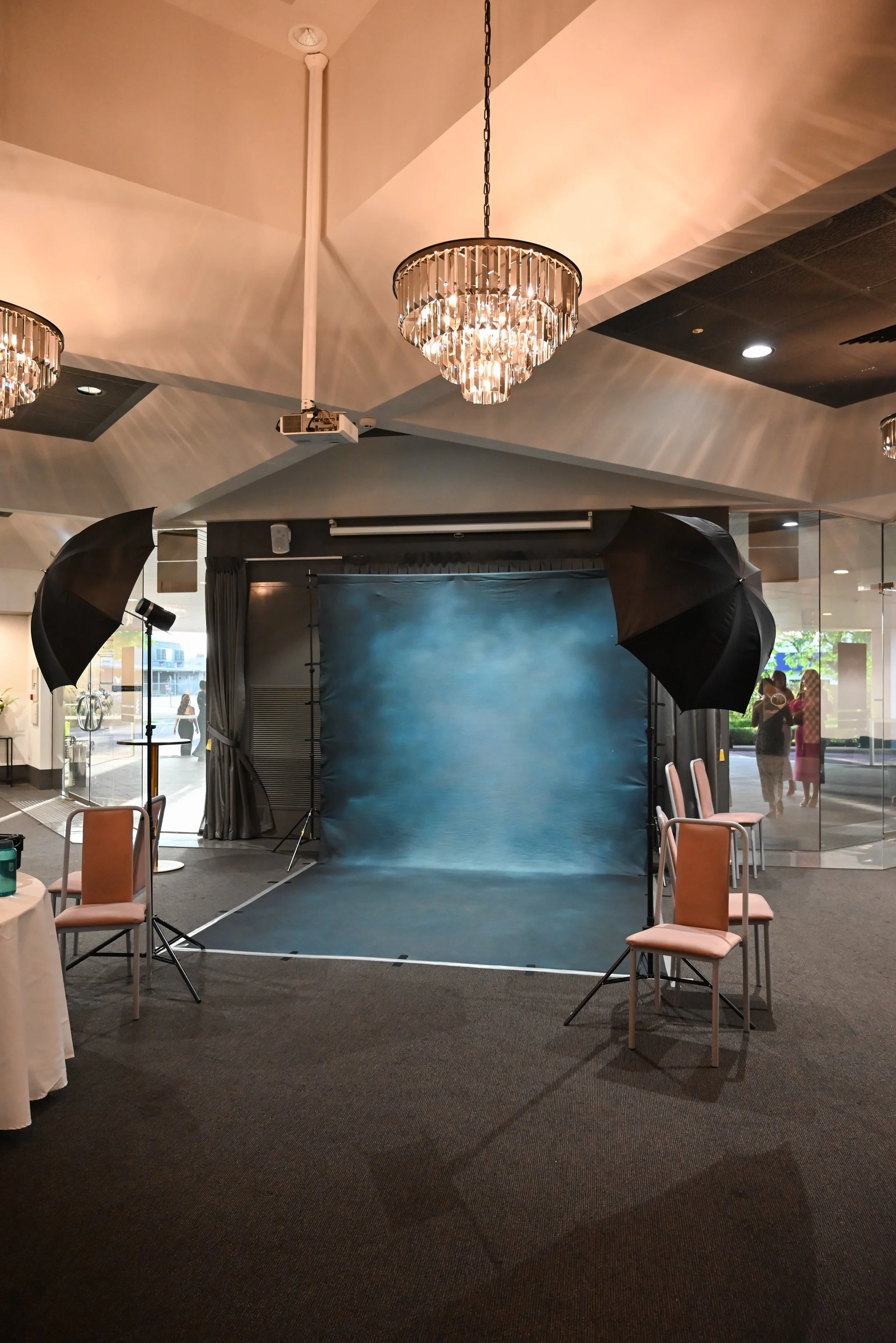 Indoor photography studio setup with a blue backdrop, surrounding lighting equipment, chairs, and people visible through glass windows.